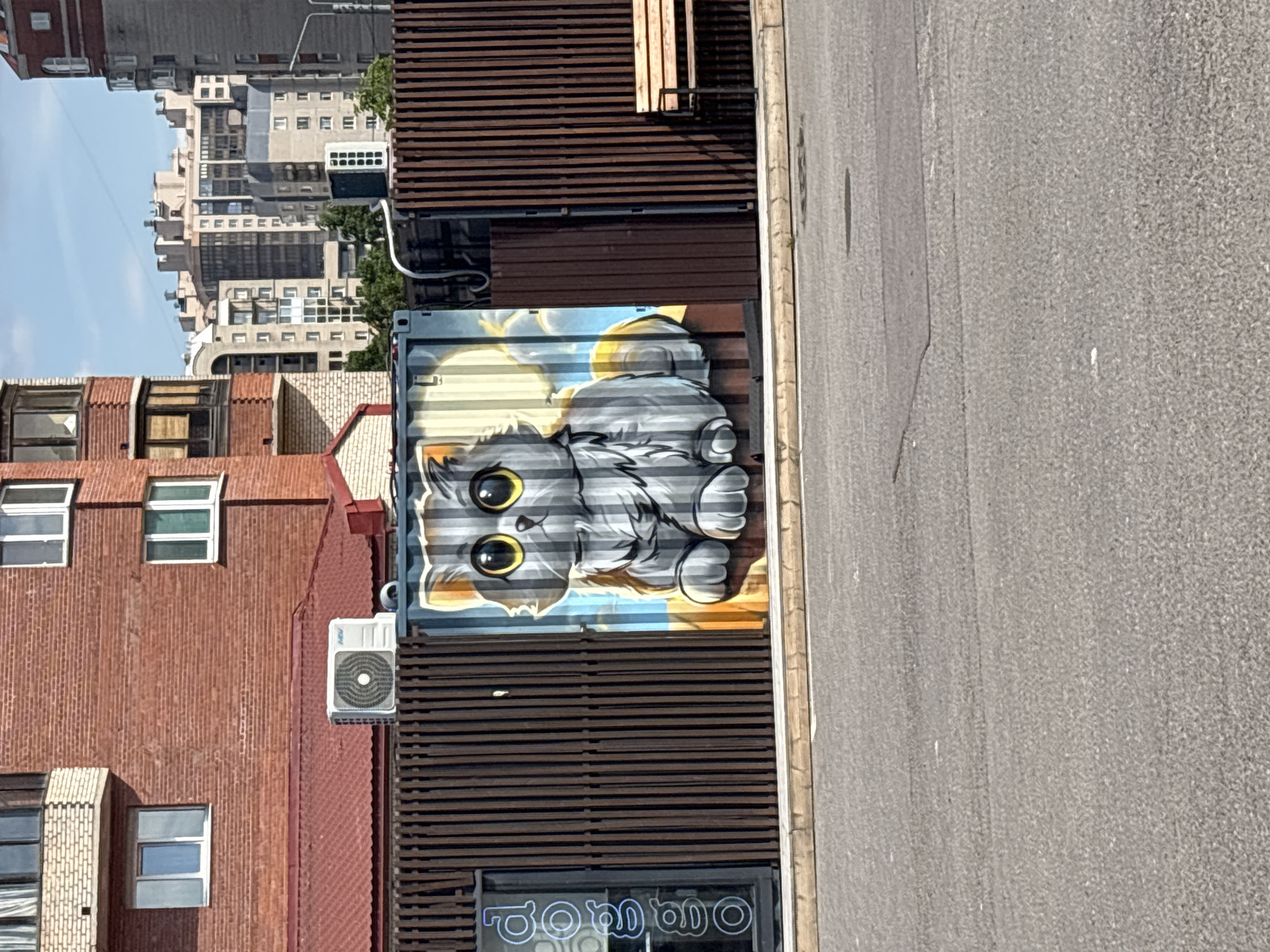 An outdoor, eye-level shot depicts an urban street scene dominated by a large, colorful mural of a cartoon cat. The mural features a fluffy, grey cat with large, bright yellow eyes and prominent paws, lying down on a background of yellow and blue clouds. It is painted on the corrugated side of what appears to be a repurposed shipping container.
To the immediate left of the cat mural is a red brick building with multiple windows, some open, and a white air conditioning unit mounted on its side. Further in the background, a cluster of various urban buildings, mostly multi-story, rise against a clear blue sky. These buildings show a mix of architectural styles and materials, including concrete and other brickwork.
Adjacent to and above the mural, and extending to the right, are structures made from horizontal brown slatted panels, resembling more shipping containers or modern facade elements. An asphalt road or paved area runs along the right side of the image, with a curb marking its edge. The scene is brightly lit by natural sunlight, indicating daytime. Below the cat mural, partially visible on a window or sign, is stylized text that appears to read "bombo". No people or specific activities are visible.