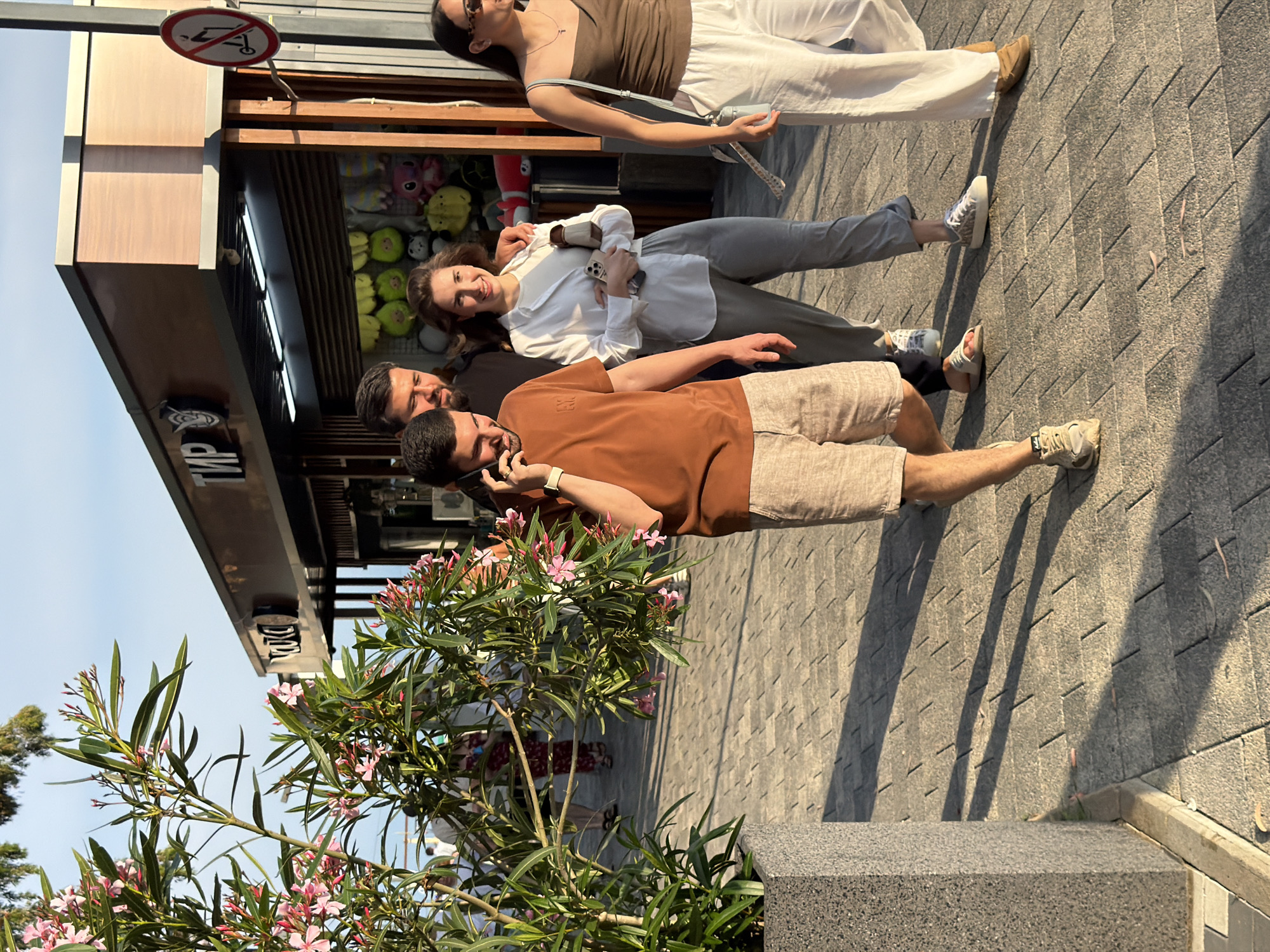 Four people are standing on a paved sidewalk in front of a shop. The group consists of two men and two women. The man in the foreground is wearing a brown t-shirt and beige shorts, and he is holding a phone to his ear. Behind him, another man is visible, wearing a dark shirt. To their right, a woman in a white shirt and grey pants is holding a phone. Further back, a woman in a brown tank top and white pants is also visible.
The shop behind them appears to be selling stuffed animals. A sign above the shop has a circular red and white symbol that looks like a "no entry" sign with a stroller inside. Another sign below it has Cyrillic text that includes "ТИР".
The scene is outdoors, and the lighting suggests it is daytime, likely late afternoon due to the long shadows. The weather appears to be clear and sunny. The paving stones are laid in a diagonal pattern. A flowering plant with pink blossoms is in the foreground, to the left of the frame. The overall mood seems casual and relaxed, with the individuals appearing to be enjoying a pleasant day.