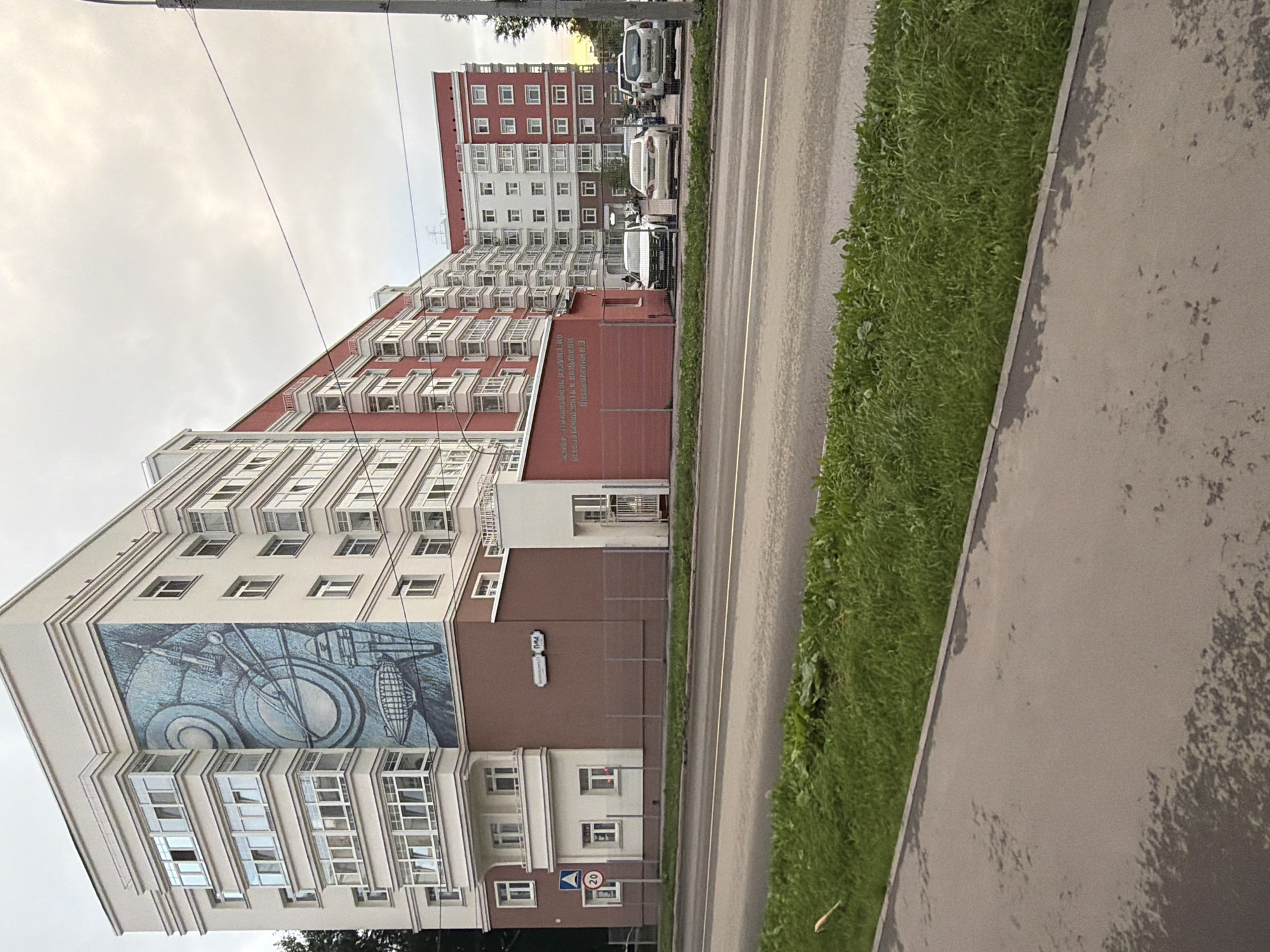 The image shows a tall, multi-story apartment building in Perm, Russia. The building has a distinct architectural style with many balconies and large windows. A mural is painted on the side of the building, depicting what appears to be an abstract design with geometric shapes and possibly industrial elements. In front of the building, there is a sidewalk, a grassy area, and a road. Several cars are parked along the road, including white vans and a dark-colored car. The sky is overcast, suggesting a cloudy or possibly rainy day. The scene appears to be an urban residential area. There is some visible text on a sign on the building, which is in Russian. The overall impression is that of a typical day in a Russian city, with everyday objects and infrastructure present.
