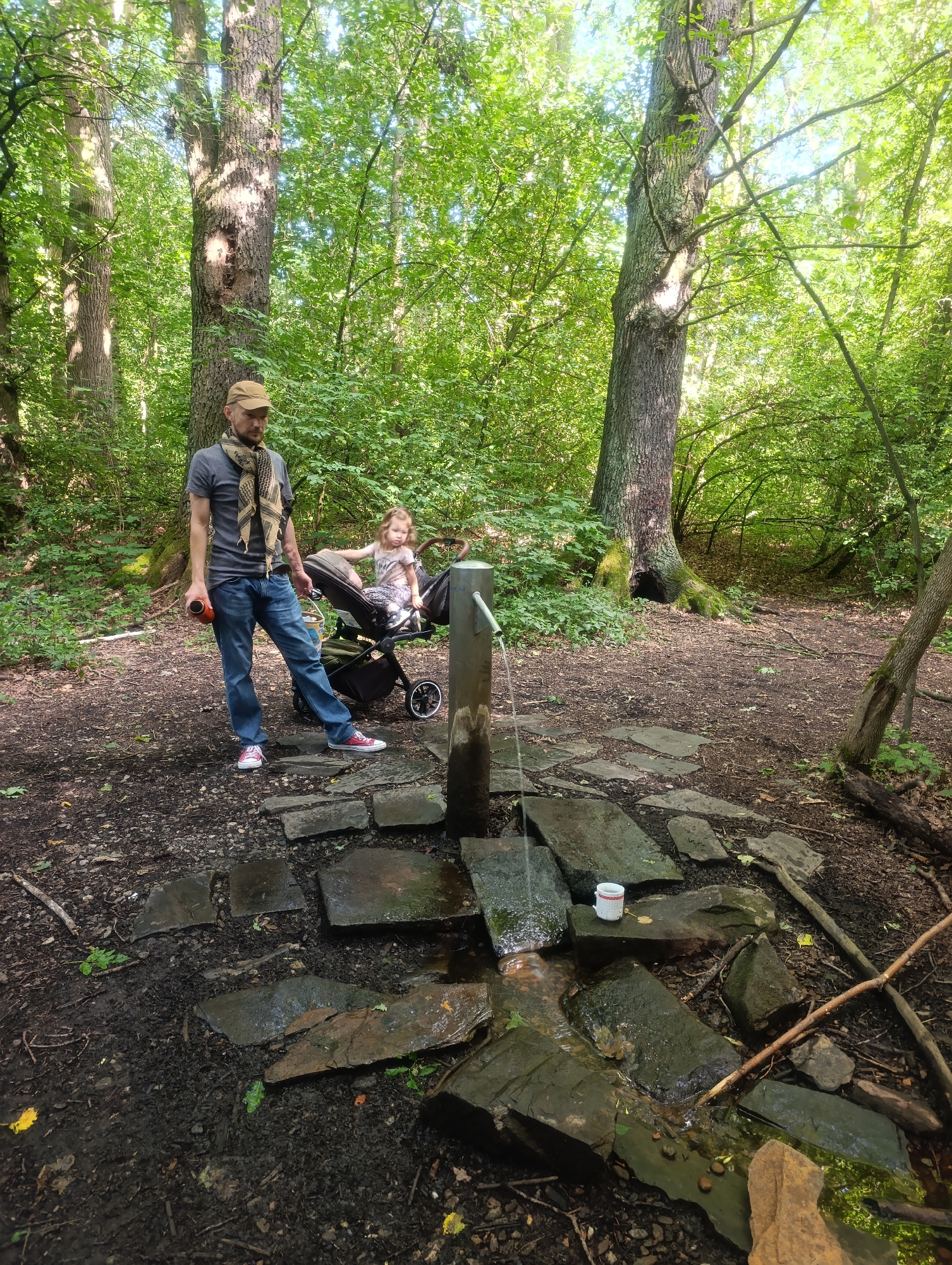 A man stands near a natural spring in a forest, holding an orange bottle. He is wearing a grey t-shirt, blue jeans, a patterned scarf, and a beige cap. In the background, a young child sits in a stroller. The spring flows over flat stones, with a white cup resting on one of them. The setting is a wooded area with tall trees and lush green foliage. This natural spring is located in the city of Olomouc, Czechia, specifically within the Svatý Kopeček nature park.