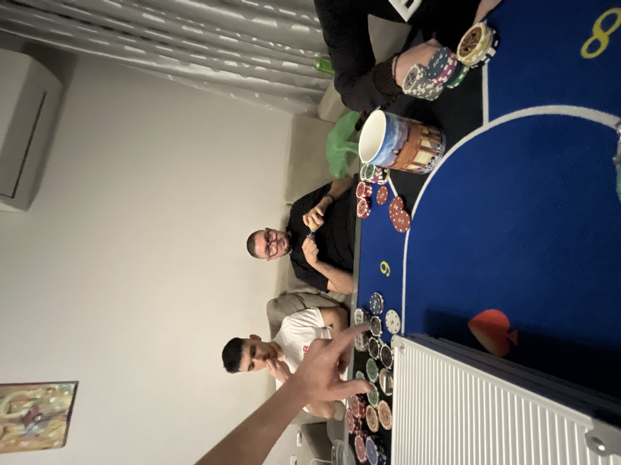 Three people are sitting around a blue poker table with chips and cards visible. One person is wearing a white t-shirt and another is wearing a black t-shirt. A third person is partially visible at the top. The setting appears to be indoors in a living room. There is a painting hanging on the wall. The location is Burgas, Bulgaria.