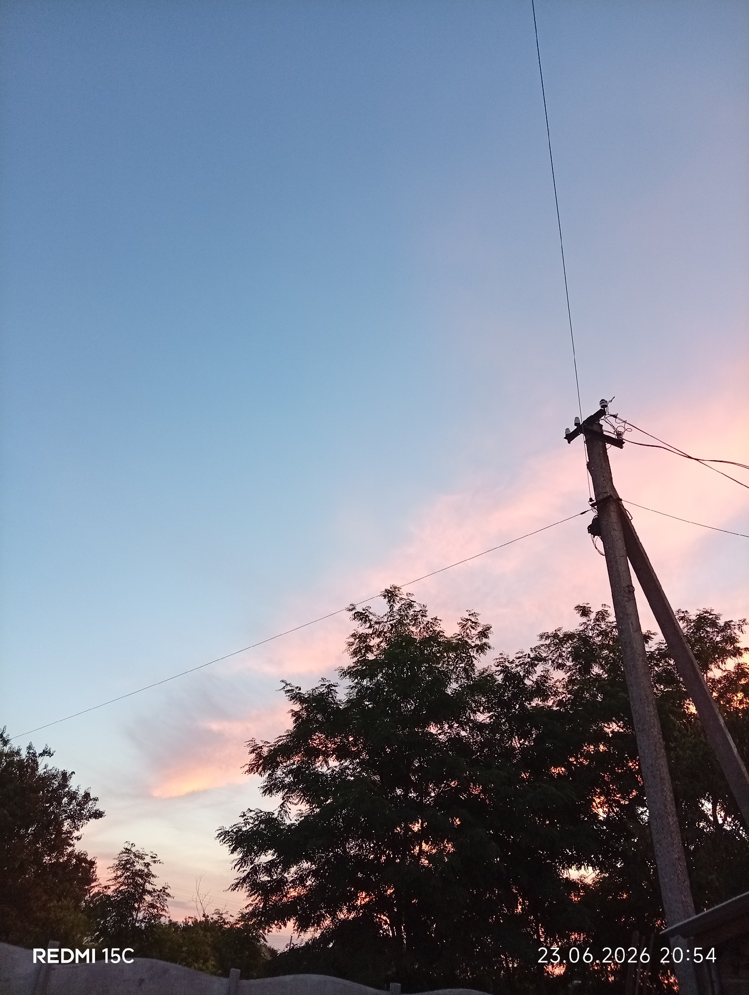 This is a vertical, medium-shot photograph taken at dusk. The scene depicts a rural or suburban setting with a clear, pale blue sky transitioning to soft pink and orange hues near the horizon, indicating the end of the day. Silhouetted against the colorful sky are the dark green leaves of several trees, some appearing dense and full, while others are sparser.
Dominating the right side of the image is a concrete utility pole with diagonal bracing. Wires run from the top of the pole, disappearing out of frame and also stretching towards the left, crisscrossing over the trees. The pole itself appears weathered and aged. In the foreground, a dark, low structure, possibly a fence or wall, runs across the bottom of the frame.
The overall mood is tranquil and serene, characteristic of a peaceful sunset. The natural beauty of the sky is contrasted with the man-made utility infrastructure.
Visible text at the bottom left corner reads "REDMI 15C". At the bottom right corner, a date and time stamp reads "23.06.2026 20:54". The location context provided is Mykolaivka, Ukraine.