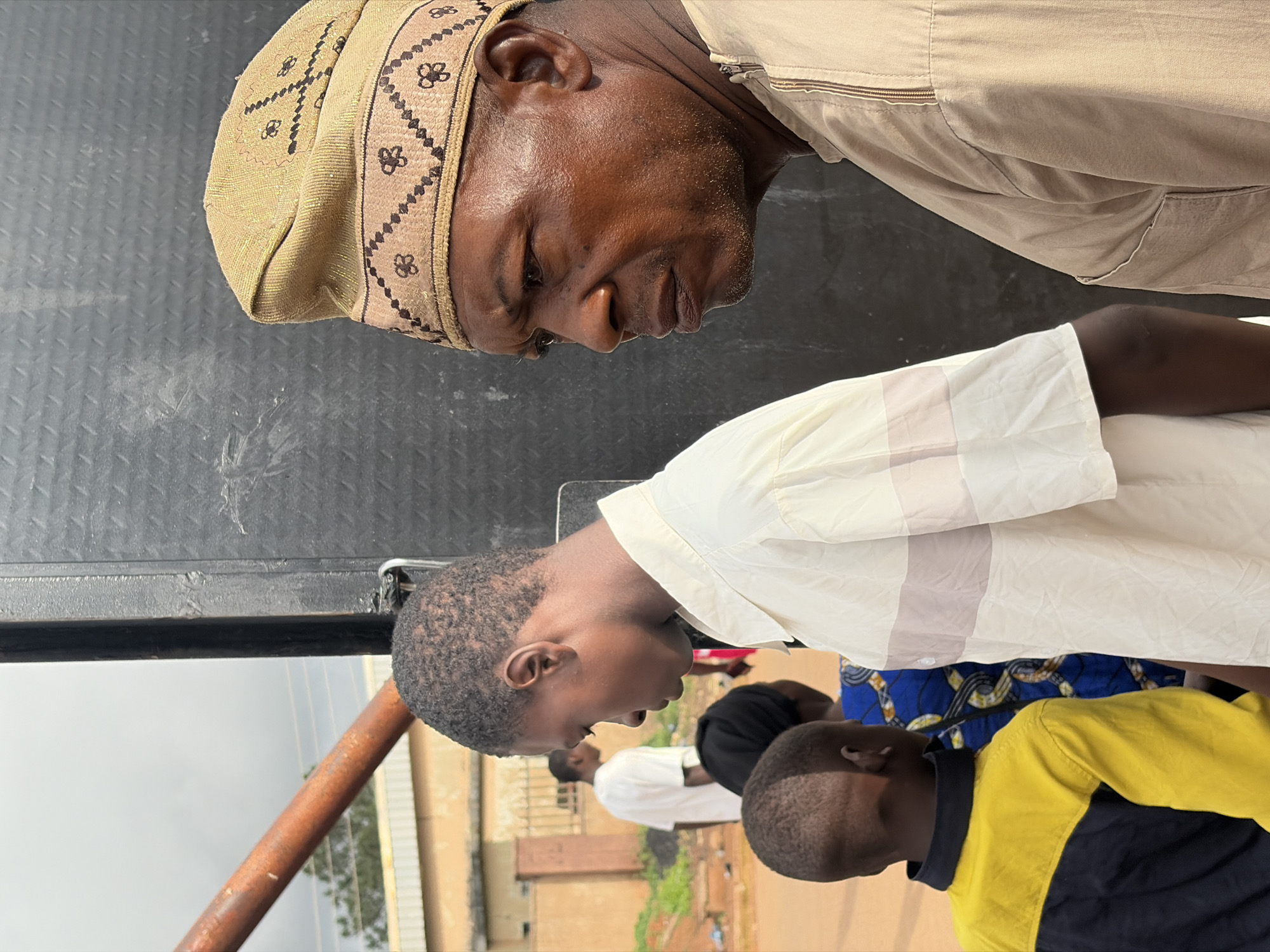 The image depicts a group of people standing in front of a dark, textured surface, possibly a large speaker or a wall. The main subjects are two men and a young boy. The man on the right, in the foreground, is wearing a light brown traditional cap with a patterned band and a matching light brown outfit with a zipper detail at the neck. He is looking to his left with a serious expression. To his left, a young man is visible from the side, wearing a cream-colored shirt with horizontal stripes. Further left, a young boy is seen from the back, wearing a bright yellow and dark blue jacket. In the background, other individuals and buildings are visible, suggesting an outdoor urban or semi-urban setting. The environment appears to be dusty with some vegetation. The overall lighting suggests it is daytime. The location is identified as Jos, Nigeria.