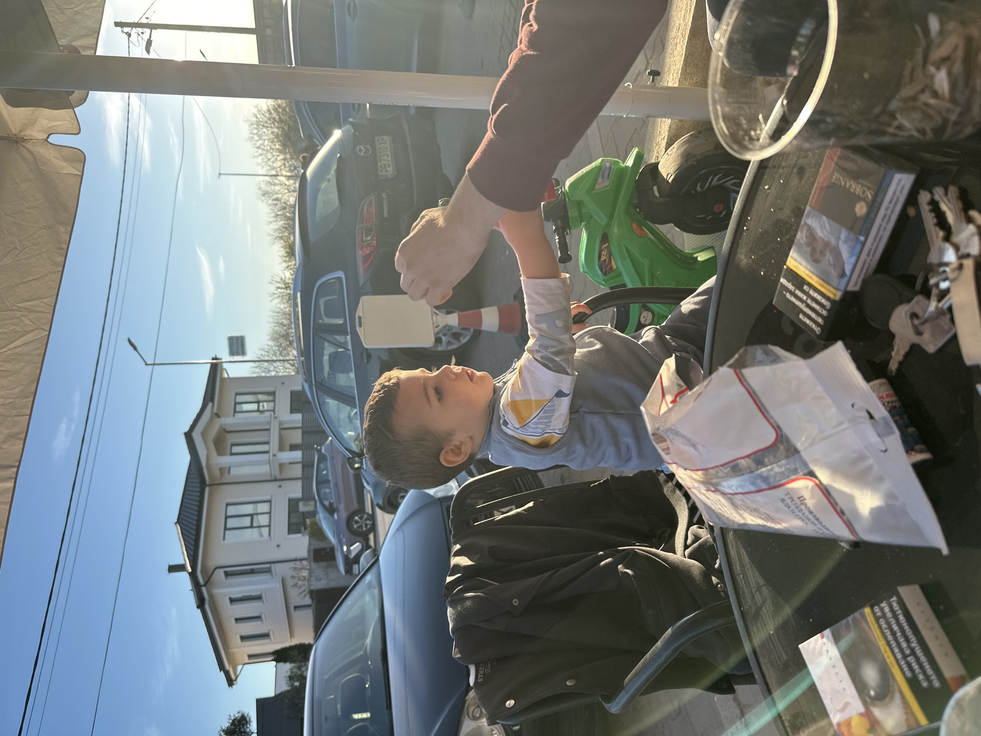 The image shows a child seated on a green toy vehicle. A hand holds up a white, rectangular object, possibly a card or piece of paper, near the child's face. The scene appears to be outdoors, possibly at a cafe or outdoor seating area. Visible on the table are keys, a pack of Sobranie cigarettes, and a white plastic bag. Cars and a multi-story building are visible in the background. The license plate of a vehicle shows "PB7310 XP", which may indicate the city of Plovdiv, Bulgaria. The environment suggests a sunny day in an urban setting.