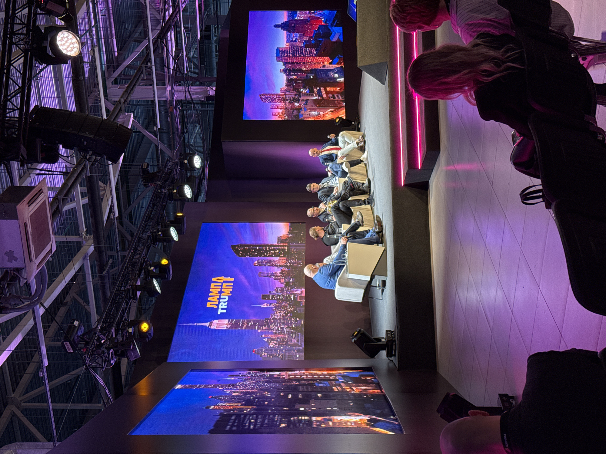 The image captures a scene at the "Lampa Trump" event in Perm, Russia. A panel discussion is taking place on a stage. Several men are seated on chairs on the stage, facing the audience. Behind them are three large screens displaying cityscape images. The screens are illuminated with a blue and purple hue. The overall lighting of the scene is a mix of bright white spotlights and neon purple accents. The venue appears to be a large indoor space, with exposed beams and rigging visible in the ceiling. In the foreground, an audience member is visible. Their face is partially obscured, but they seem to be holding a camera or phone, possibly recording the event.