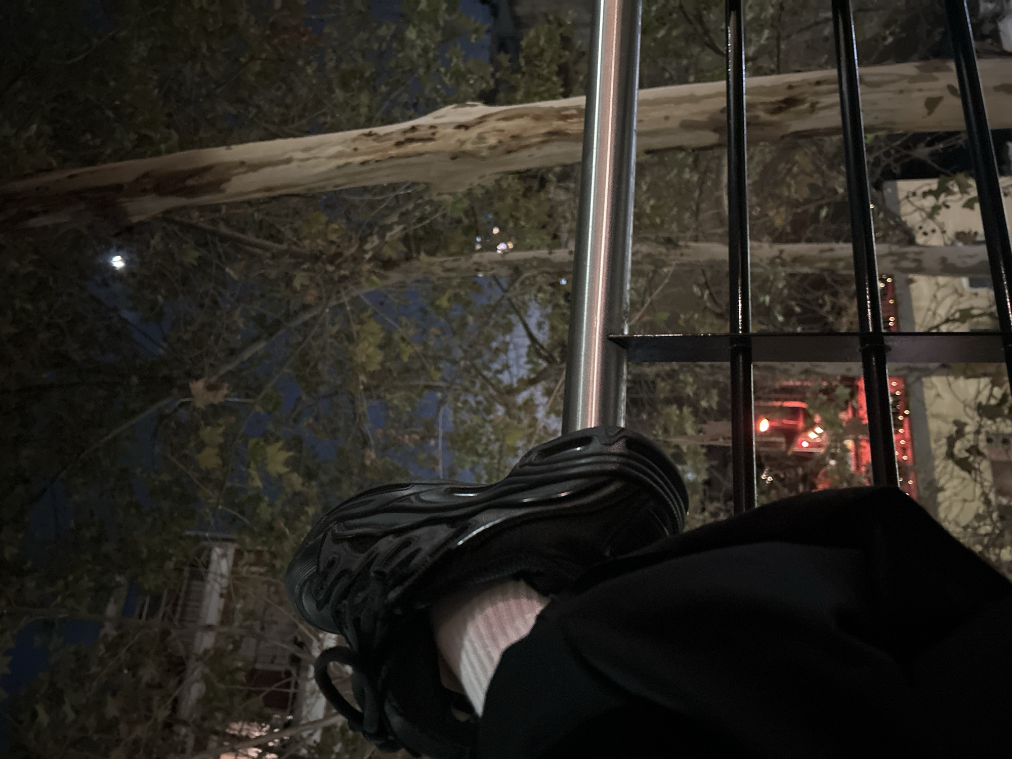 The image shows a person's leg resting on a metal pole in an outdoor setting, likely a cafe or restaurant. The person is wearing a black shoe with a thick sole, a white sock, and black pants. The background includes trees, some with light-colored bark, and buildings visible through the branches and a black metal fence. There are also hints of city lights in the distance. Given the image's location in Buenos Aires, Argentina, the presence of outdoor cafes and the city's architecture, the scene may be a street in a neighborhood known for its nightlife.