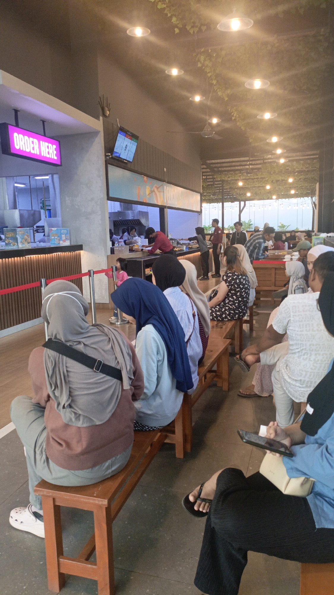 The image captures a busy scene inside a restaurant or food court, possibly in Pelaihari, Indonesia, with people waiting in line. A prominent illuminated "ORDER HERE" sign is visible on the left. Several individuals are seated on wooden benches, facing the counter area where staff are preparing orders. Many patrons are wearing headscarves, suggesting a cultural context. The environment appears to be an indoor dining space with overhead lighting and decorative foliage. The overall atmosphere suggests a popular dining establishment with a steady flow of customers.