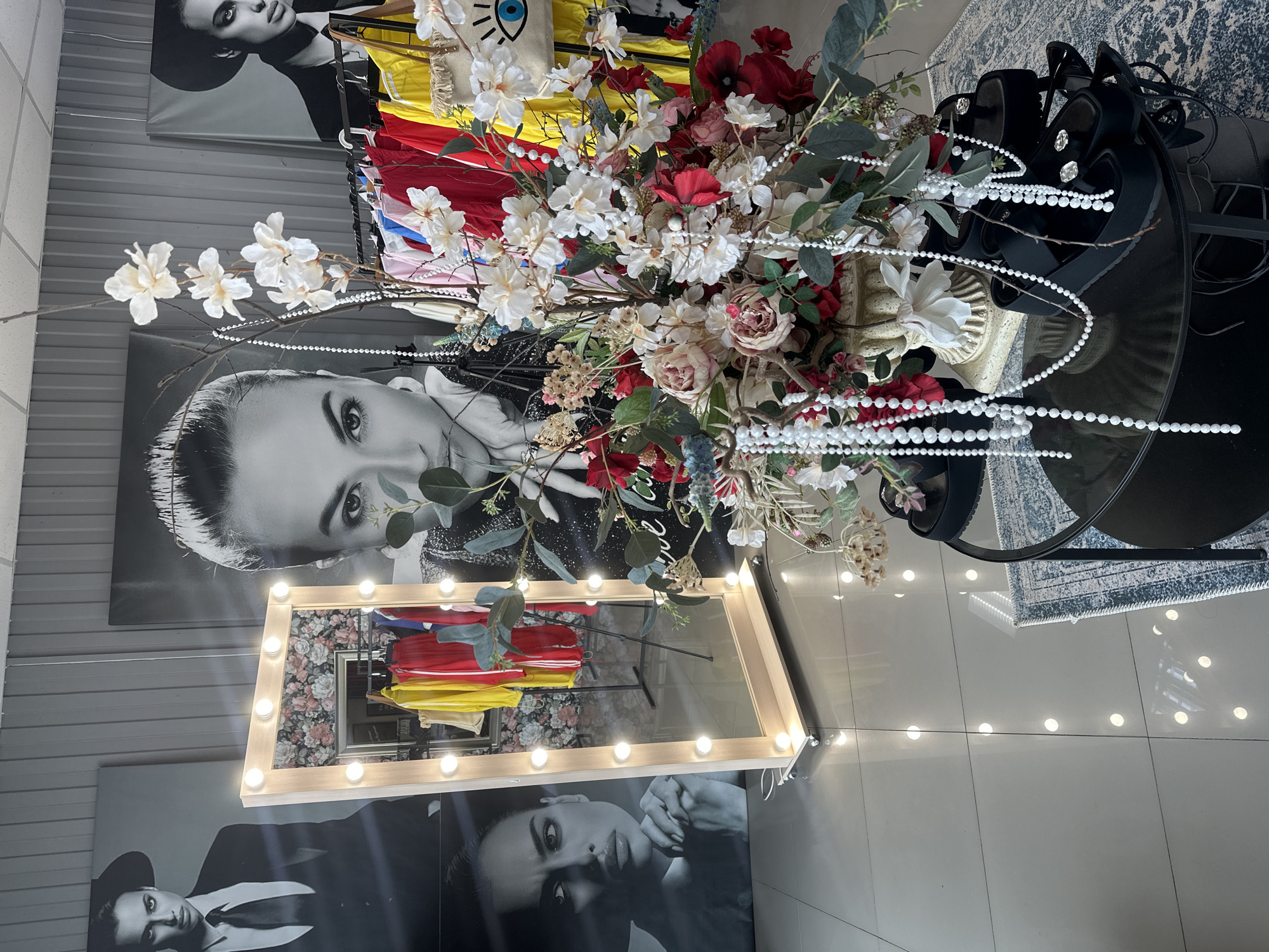 The image shows the interior of a retail establishment, likely a boutique, located in Votkinsk, Russia. The space is decorated with large black and white photographs of models displayed on a gray, paneled wall. In the foreground, a lighted vanity mirror with a wooden frame stands on wheels. To the right of the mirror, a clothing rack displays brightly colored garments, including red, yellow, and blue items. A large floral arrangement, featuring red and white flowers, is placed in a decorative urn on a black, round table. Black platform sandals with decorative elements are arranged on the table. The floor is tiled and highly reflective, with the lights from the mirror and overhead fixtures creating numerous reflections. The overall impression is of a stylish and modern retail environment.