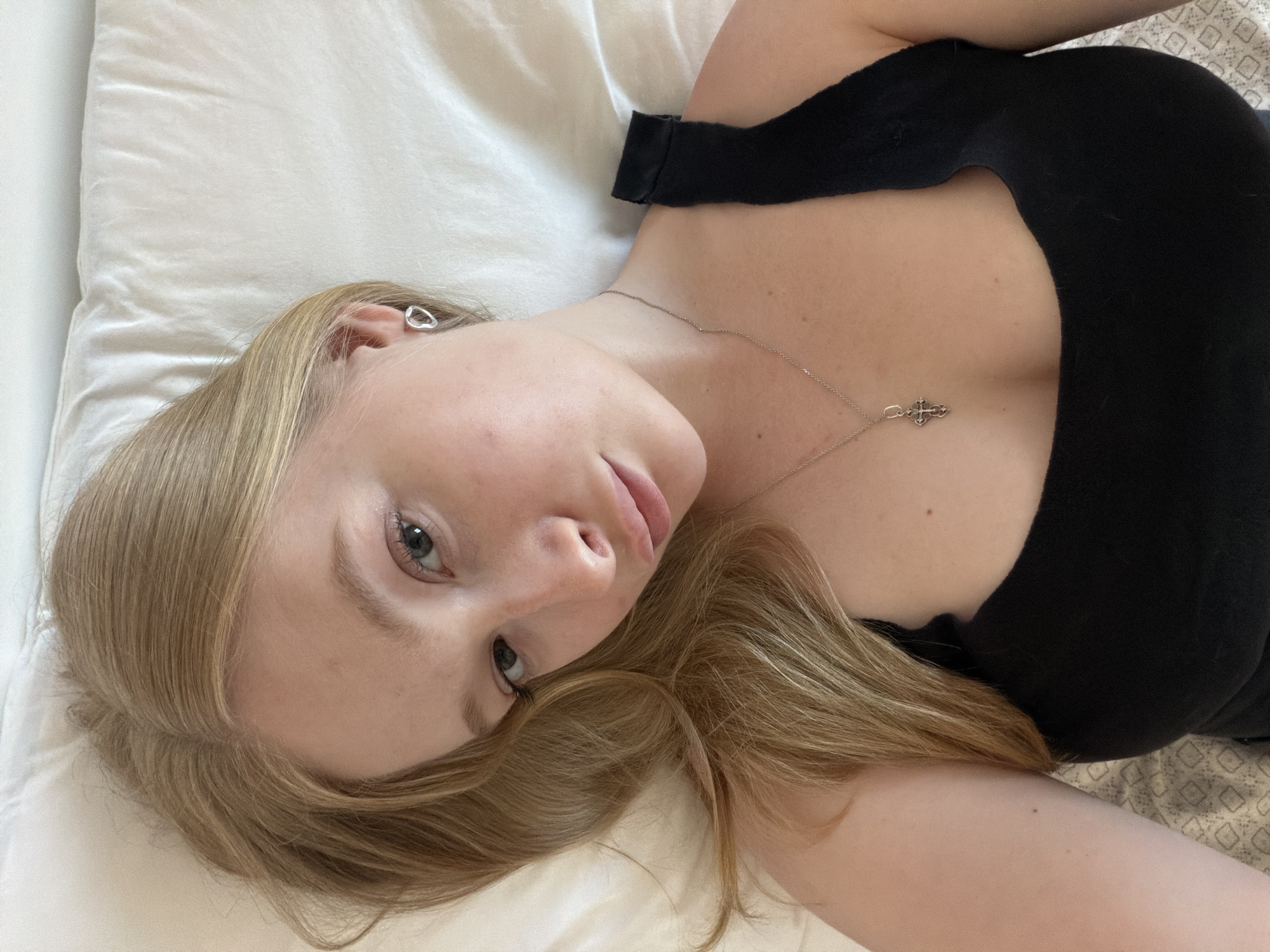 A young woman with blonde hair lies on a white bed, looking up at the camera. She is wearing a black tank top and a silver necklace with a cross pendant. She also has a silver earring in her left ear. The background is a white, wrinkled sheet. The lighting is soft and natural, coming from above. The image appears to be a selfie, taken from a high angle. Rossiyskiy, Russia is not depicted in this image.