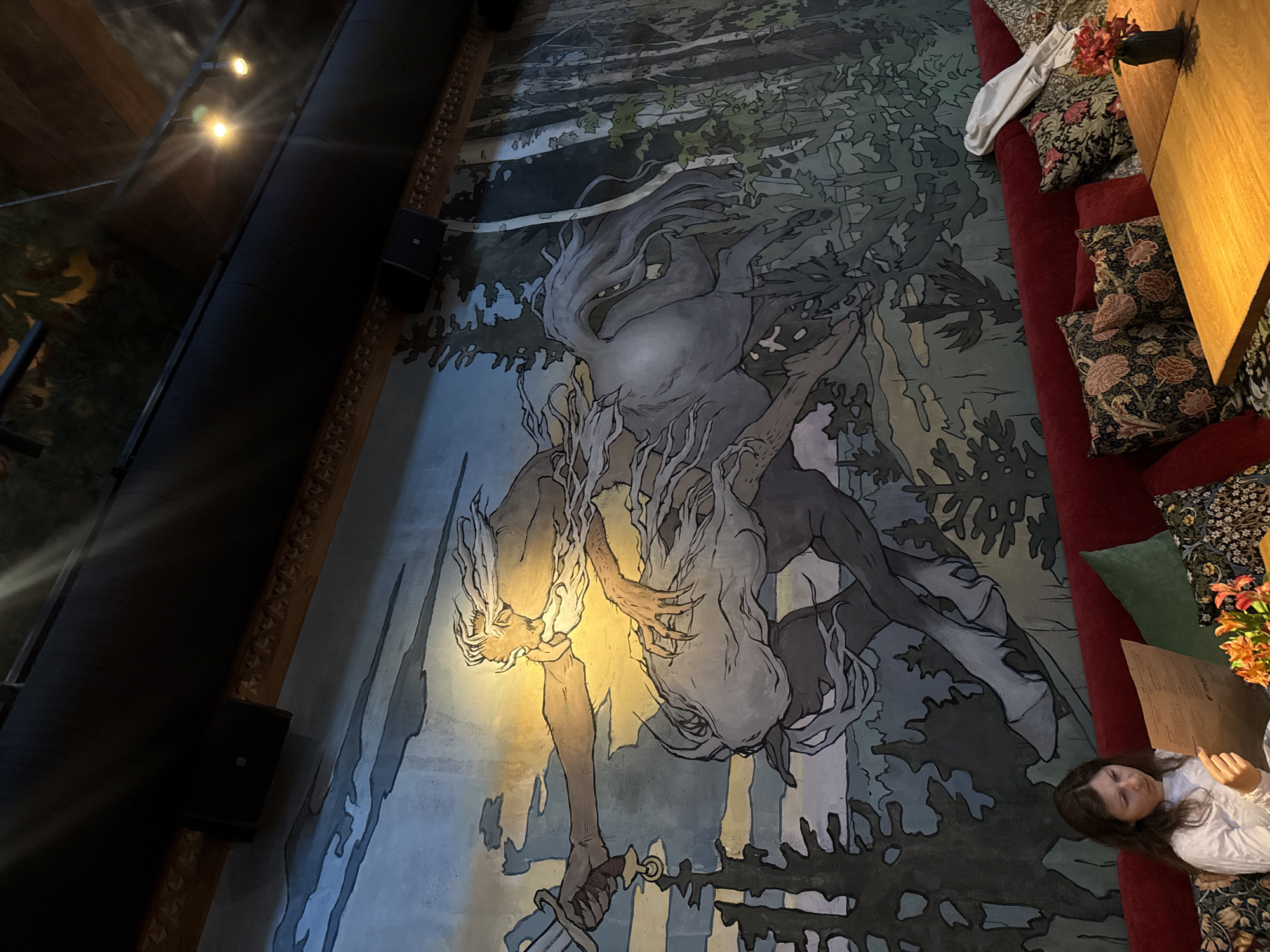 A woman with long brown hair, wearing a white long-sleeved shirt, sits on a red couch in a restaurant, reading a menu. She is in Moscow, Russia. The wall behind her is adorned with a large mural depicting a mythological scene. The mural features a centaur-like creature with flowing white hair and beard, wielding a sword. The background of the mural shows a forest scene with trees and a dramatic sky, illuminated by a bright yellow light source representing the sun. The couch is decorated with patterned pillows. A wooden table is visible in the foreground with a small vase of flowers and a bottle. The ceiling features industrial elements like pipes and lighting fixtures.