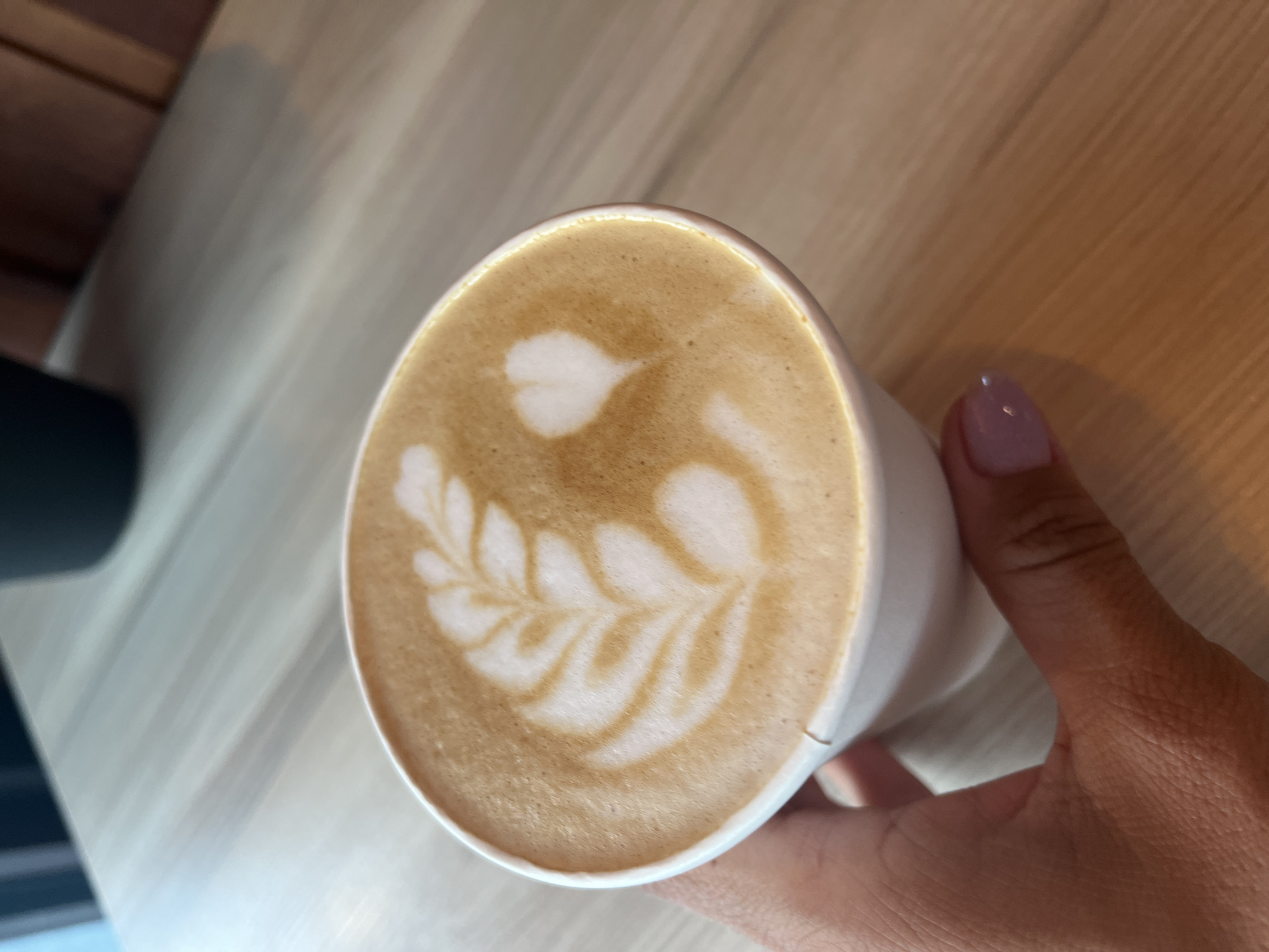 A hand with light pink nail polish is holding a white paper cup filled with coffee. The coffee has latte art on top, with a fern-like pattern and a heart shape made from milk foam. The cup is resting on a light-colored wooden surface. The background is slightly blurred, showing a dark object and more of the wooden table. The image appears to be taken indoors, possibly in a cafe. There is no information in the image to suggest the location is Petropavlovsk-Kamchatsky, Russia.