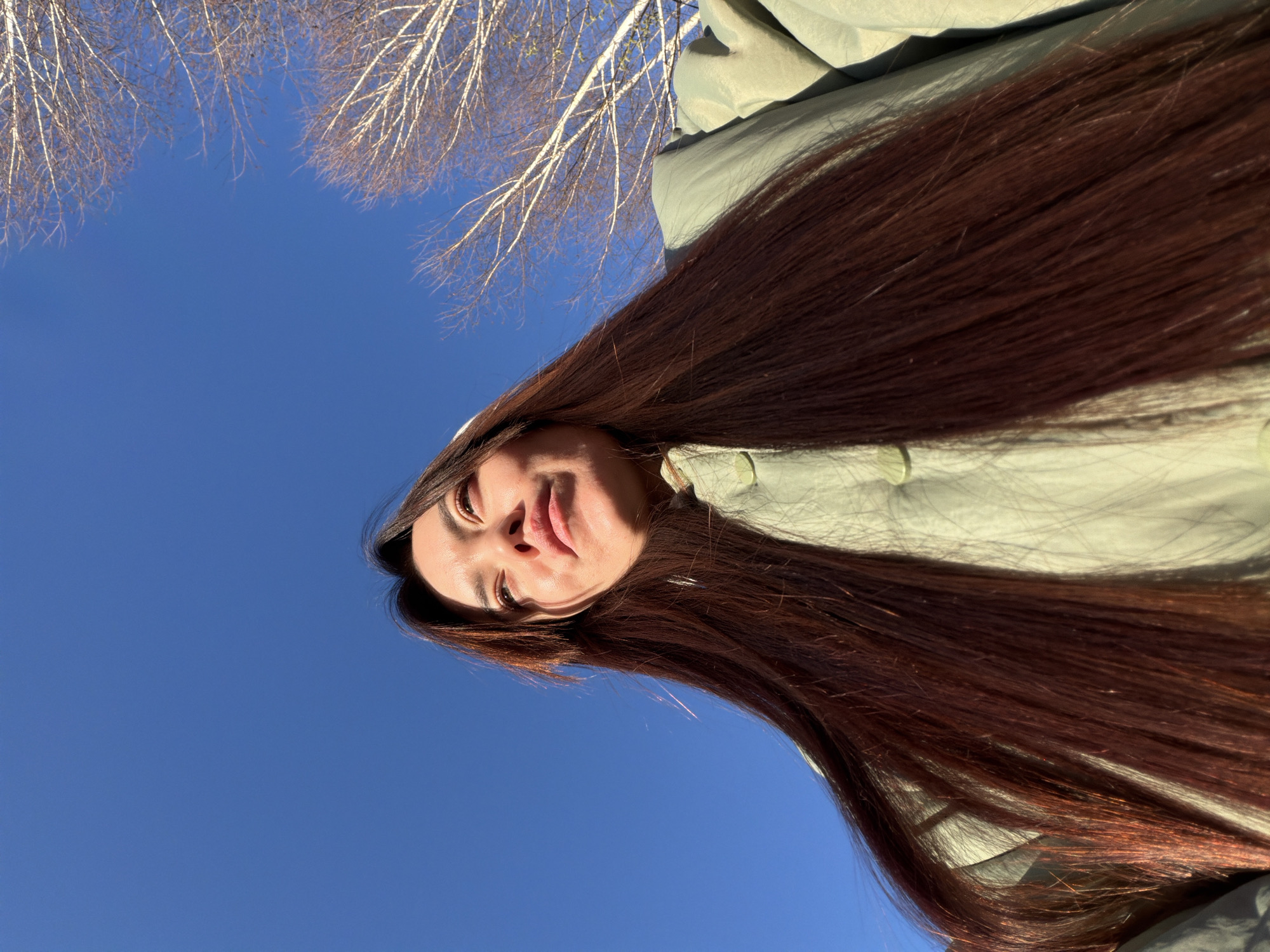 The image shows a woman with long, auburn hair wearing a light green jacket, lying on her back looking up at the sky. The sky is clear blue with bare tree branches visible in the upper portion of the frame. The location is Chaikovsky, Russia. The specific activity is the woman observing her surroundings.