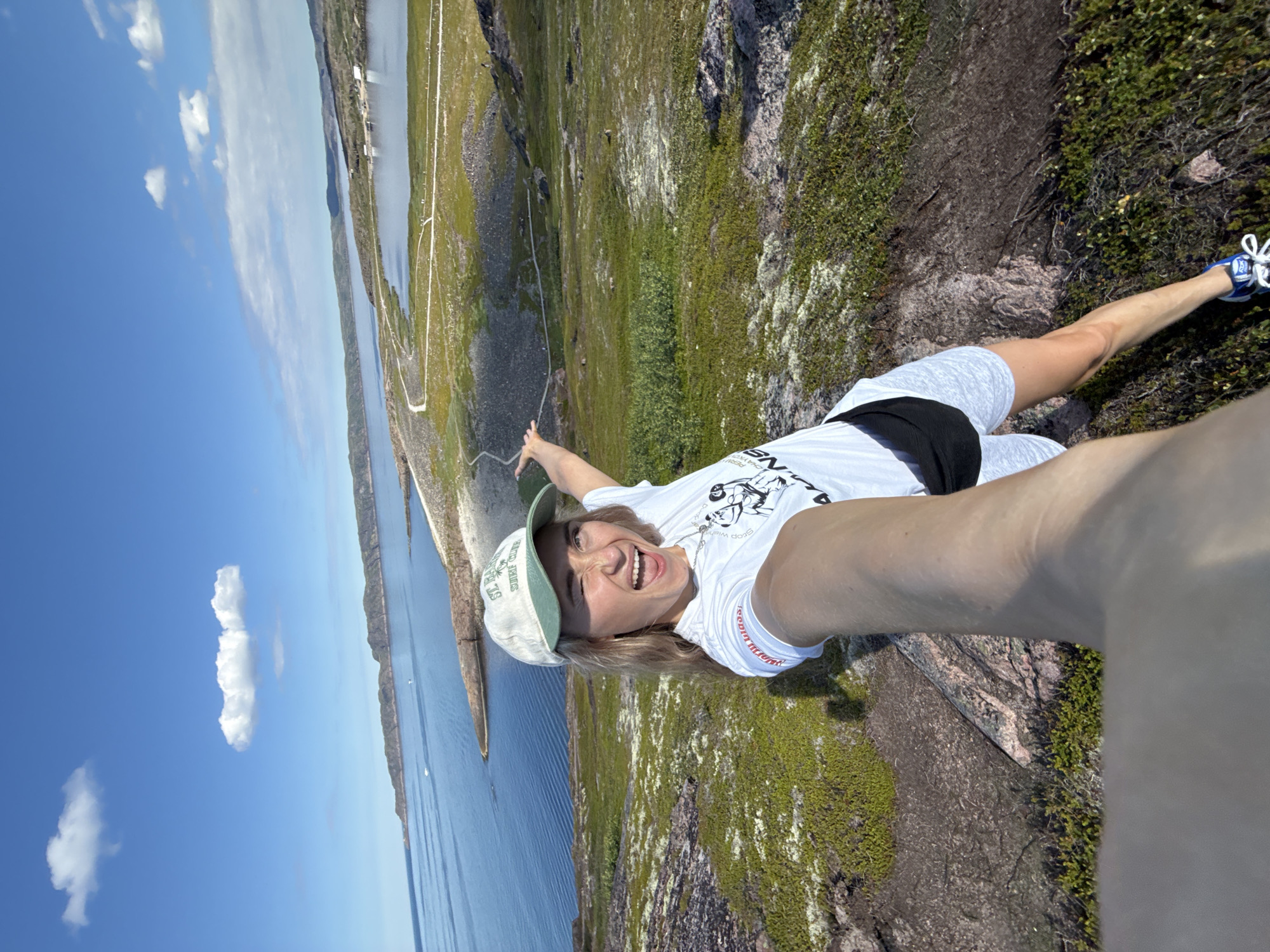 A woman takes a selfie on a rocky, moss-covered cliff overlooking a body of water. She is wearing a white t-shirt with a graphic print, grey shorts with a black waistband, a light-colored baseball cap with green trim, and blue and white athletic shoes. Her right arm is extended, holding the camera. She is smiling broadly at the camera. The background shows a vast blue sky with scattered white clouds, a long, narrow body of water, and a rocky, vegetated coastline with some structures visible in the distance. The scene suggests a daytime outdoor activity, likely hiking or exploring. The weather appears sunny and clear. The location is identified as Teriberka, Russia, a coastal village known for its natural landscapes. Visible text on the t-shirt is partially obscured, but "STOP WIND" and part of a graphic appear on the sleeve. The cap has text that is difficult to read but appears to be in English.