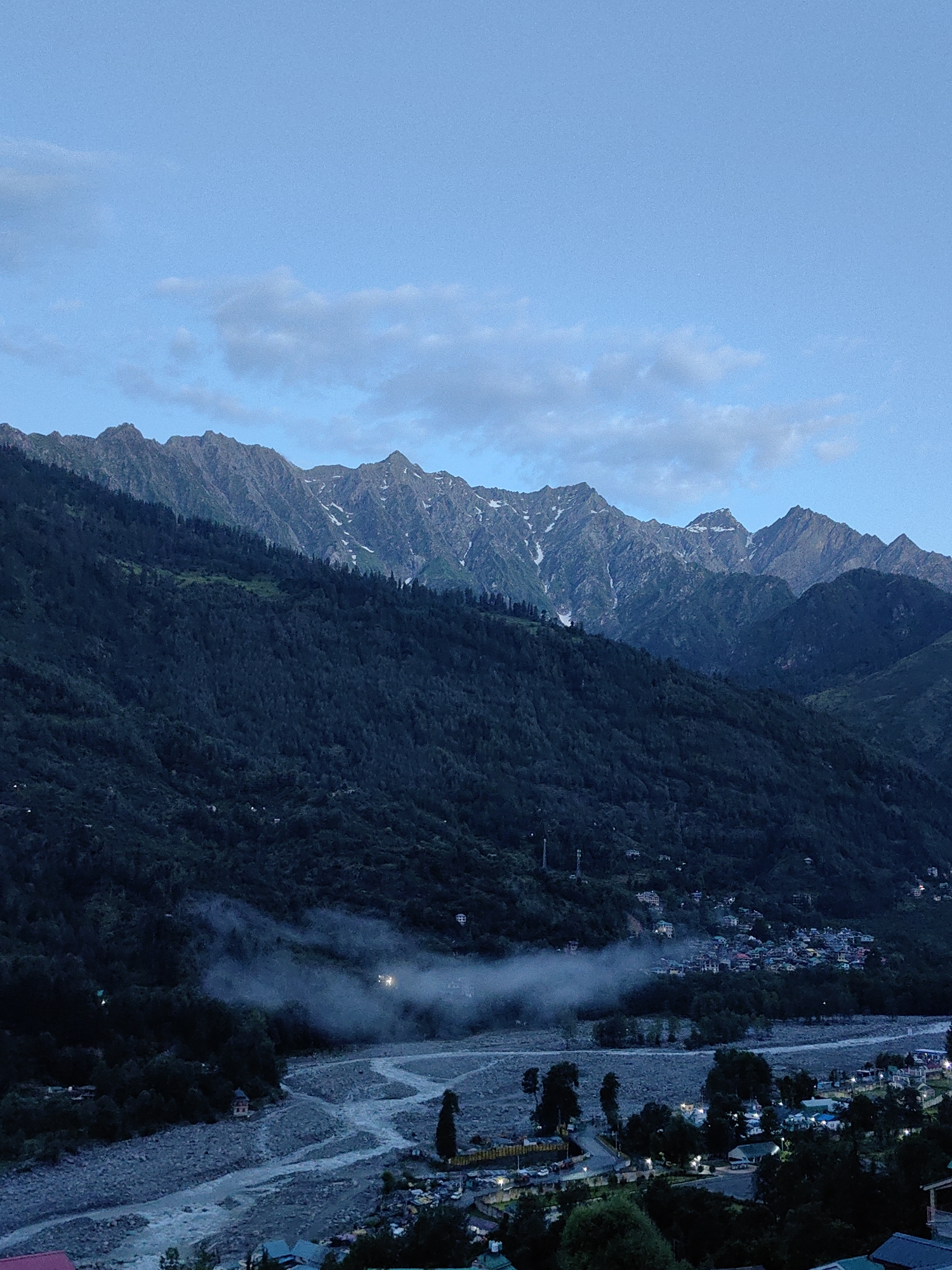 The image depicts a panoramic view of the city of Vashisht, India, at dusk. In the foreground, a wide, rocky riverbed with a river flowing through it dominates the scene. The river is bordered by a town with numerous buildings, some illuminated by lights. Trees are scattered throughout the town and along the riverbanks. In the middle ground, a dense forest covers the slopes of a mountain. A light mist hangs over the river and the lower parts of the mountain, obscuring some of the details of the town nestled within the valley. In the background, a range of majestic, snow-capped mountains rises against a clear, pale blue sky with a few wispy clouds. The overall impression is one of a serene and picturesque mountain landscape with a human settlement integrated into it.