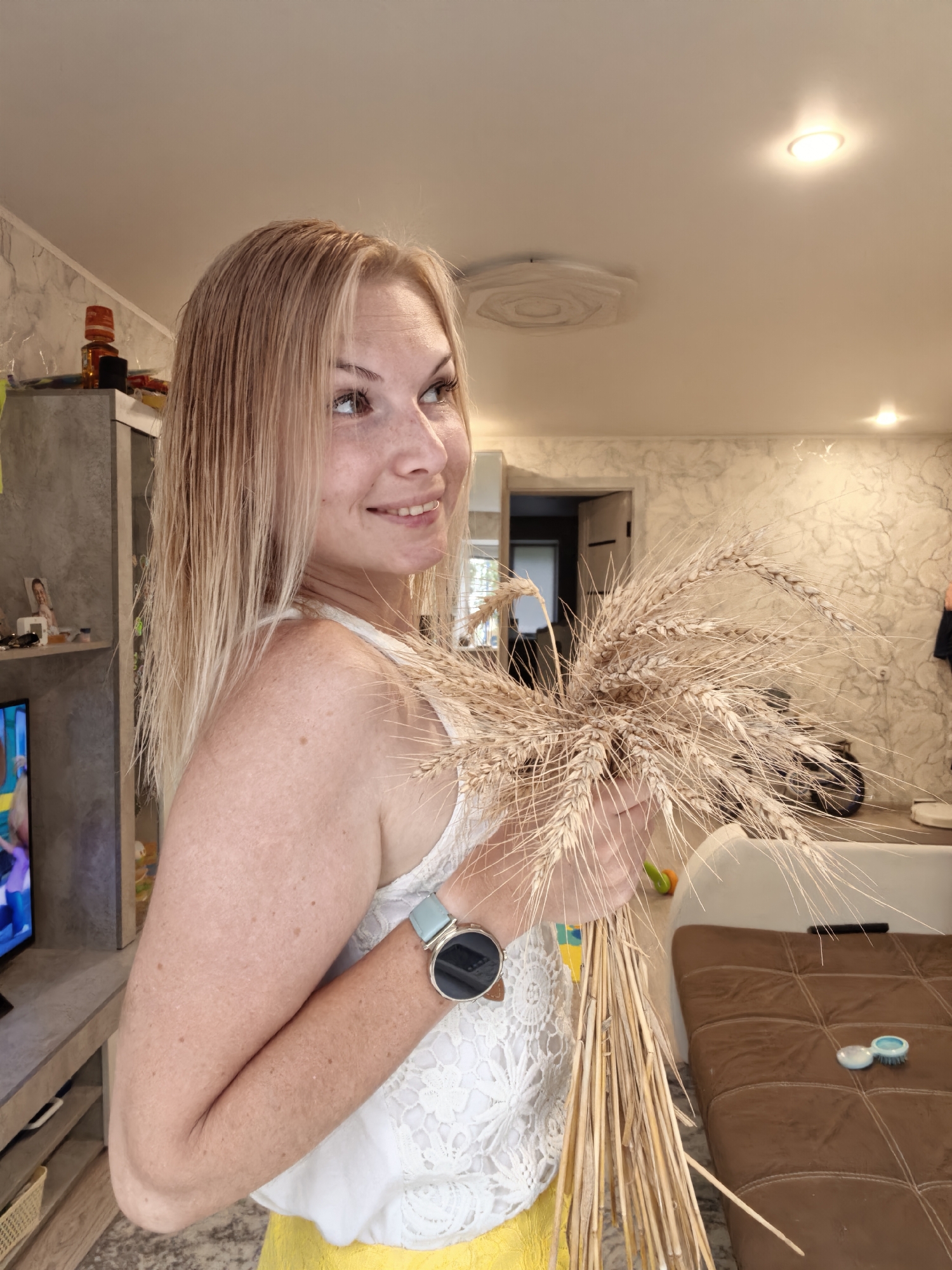 The image depicts a woman holding a bunch of wheat stalks in an indoor setting that appears to be a living room. The woman has fair skin with freckles and blonde hair. She is wearing a white top with lace details and a yellow skirt. On her wrist, she has a watch with a light blue strap. The background shows a modern interior with a couch, a cabinet, and a television. The lighting suggests it is daytime. The overall scene conveys a sense of domesticity and perhaps a celebration of harvest. The location is identified as Sterlitamak, Russia.