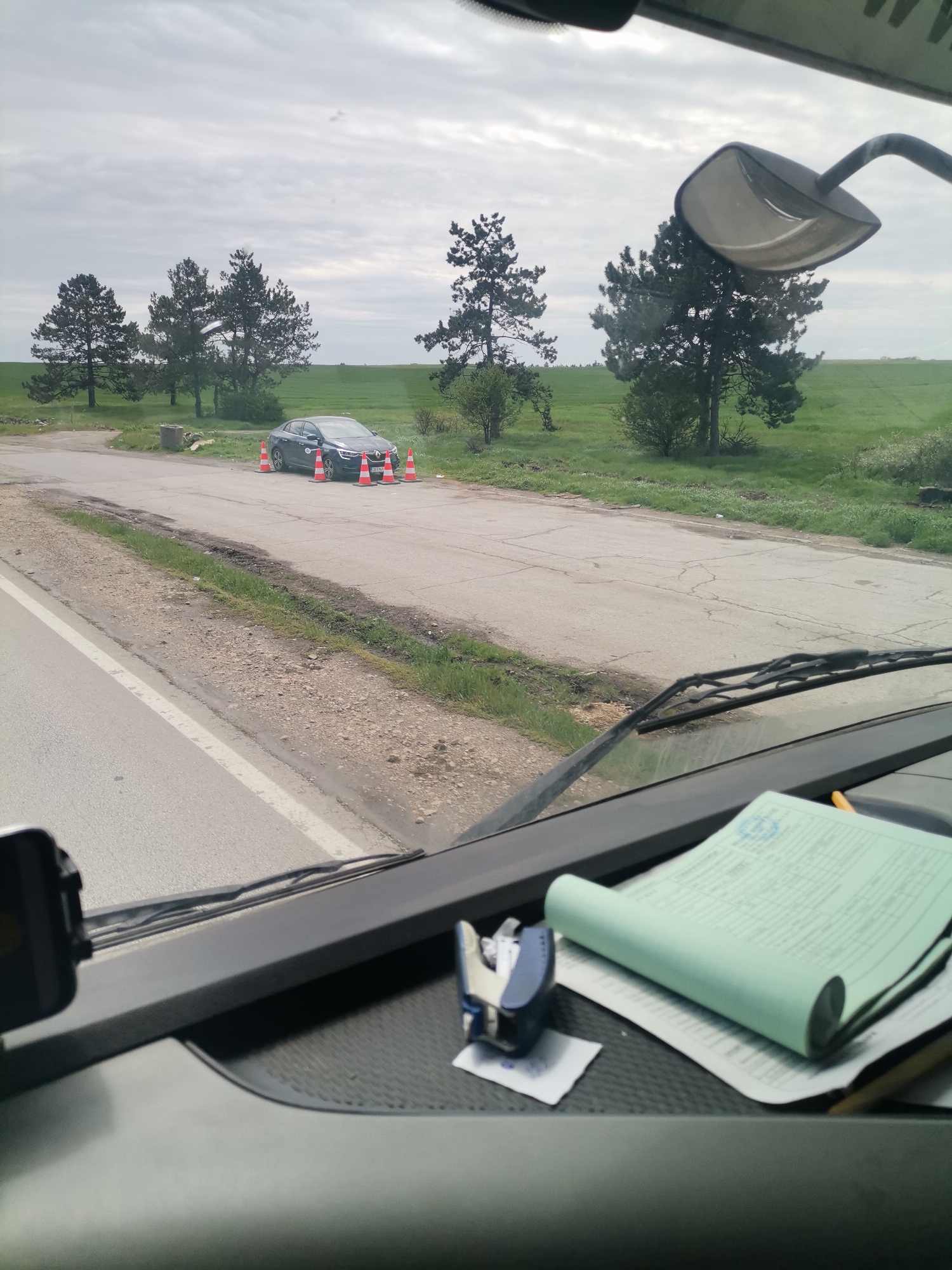 The image shows a view from inside a vehicle, likely a truck, looking out onto a road. A dark-colored car is parked on the side of the road, surrounded by orange traffic cones. Behind the car, there is a field with several trees. The sky is overcast. There is paperwork and a stapler on the dashboard of the vehicle. Based on the EXIF data, the location is Izvorsko, Bulgaria.