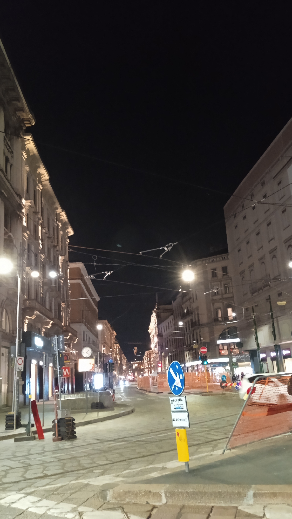 The image depicts a street scene in Milan, Italy at night. Buildings line both sides of a wide, empty street, illuminated by streetlights. The architecture appears to be of an older European style, with multiple stories and ornate facades. Overhead, a network of wires crisscrosses the sky, likely for trams or streetcars. In the foreground, there is a traffic signpost with several signs, including one with a blue circle and a white arrow indicating a right turn, and another sign that reads "ECCETTO" (except) above a smaller sign that reads "ed autorizzati" (and authorized). Construction barriers and orange netting are visible on the right side of the road. The street itself is paved with cobblestones or similar material, and tram tracks are embedded in the pavement. The overall atmosphere is one of quiet emptiness, characteristic of a city late at night.