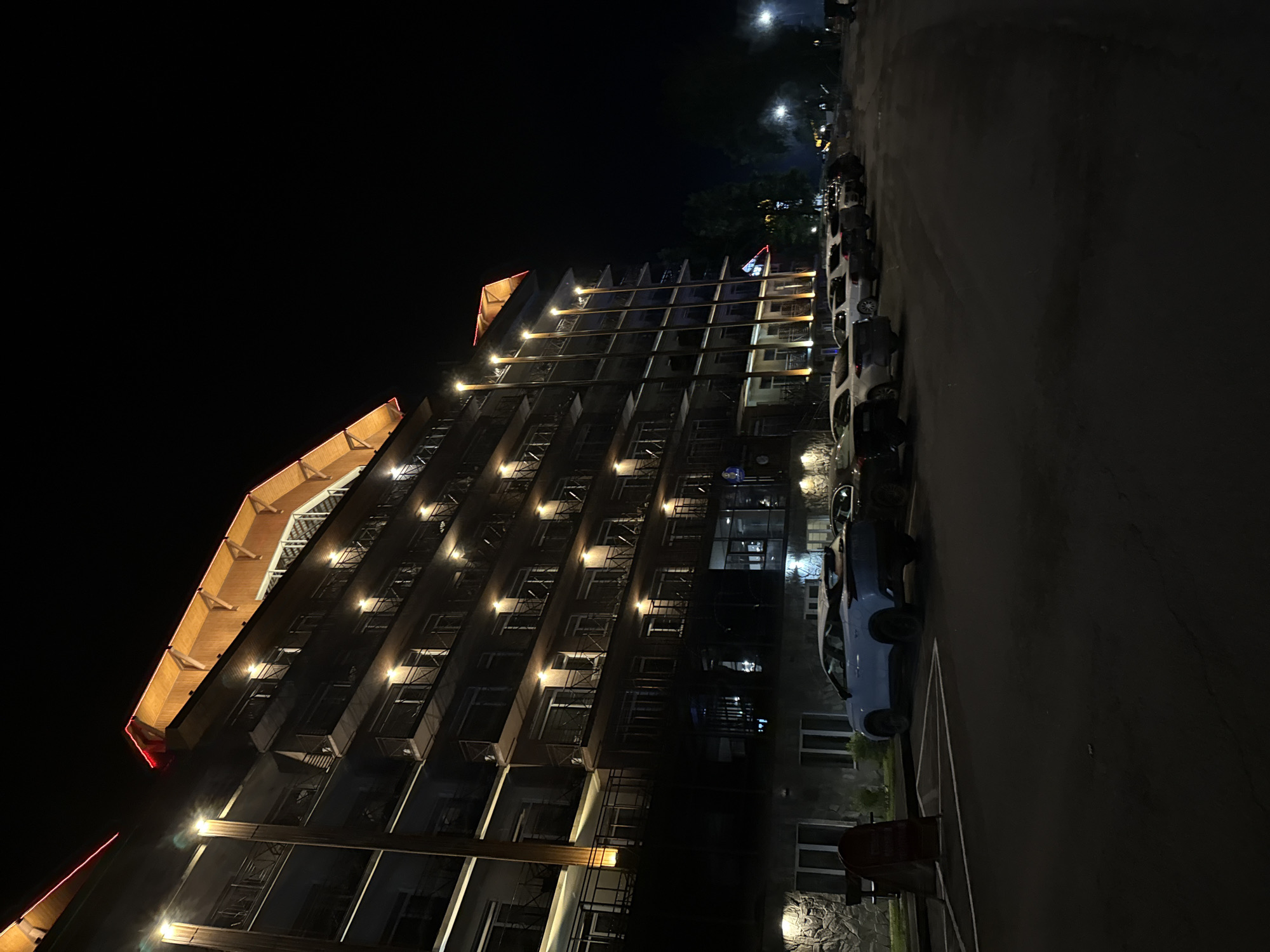 At night, a multi-story building with balconies and illuminated windows is visible on the left. The building has a wooden roof with red and yellow lighting. In front of the building is a parking lot with several cars. The ground is asphalt and there are trees visible in the background. The lighting suggests it is nighttime.