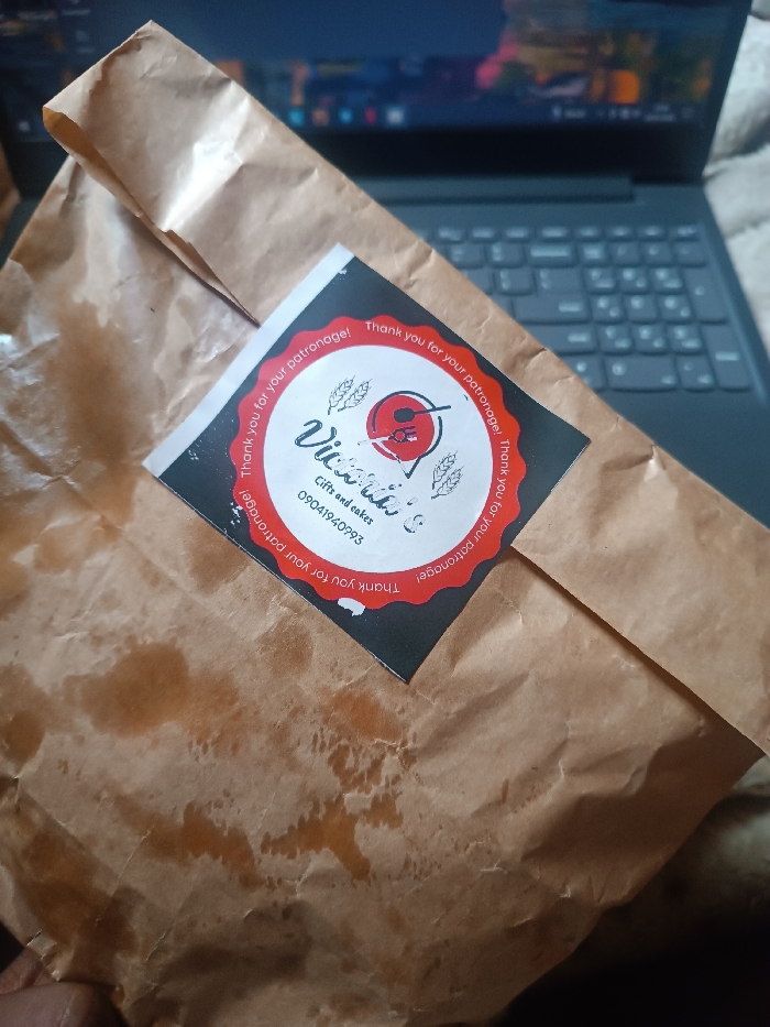 A brown paper bag, exhibiting prominent creases and grease stains, is positioned diagonally across the frame. Affixed to the bag is a circular black and red sticker. The sticker's red outer ring displays "Thank you for your patronage!" repeatedly. The black inner circle features "Vivikie's Gifts and cakes" and the Nigerian phone number "09041940993." A partial view of a human hand is visible in the bottom left corner, appearing to support the bag. In the background, a dark gray laptop with its keyboard and a blurred screen displaying a desktop environment is visible. To the right of the laptop, a light-colored, textured fabric surface is present. The scene is an indoor setting in Oye, Nigeria.