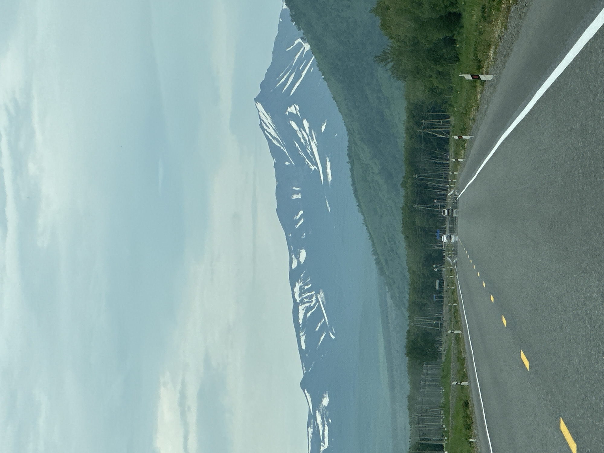 The image shows a paved road with a white dividing line and yellow dashed lines in the foreground, leading towards a vast mountain range in the background. The mountains are covered with patches of snow, indicating a high altitude or a cold climate. The sky is overcast with light gray and blue clouds. Along the sides of the road, there are utility poles and vegetation, suggesting a rural or semi-rural setting. The scene appears to be a landscape photograph taken from a moving vehicle, as evidenced by the perspective and the motion blur on the road markings. There are no people or animals visible in the image. The location is specified as Кеткино, Russia.