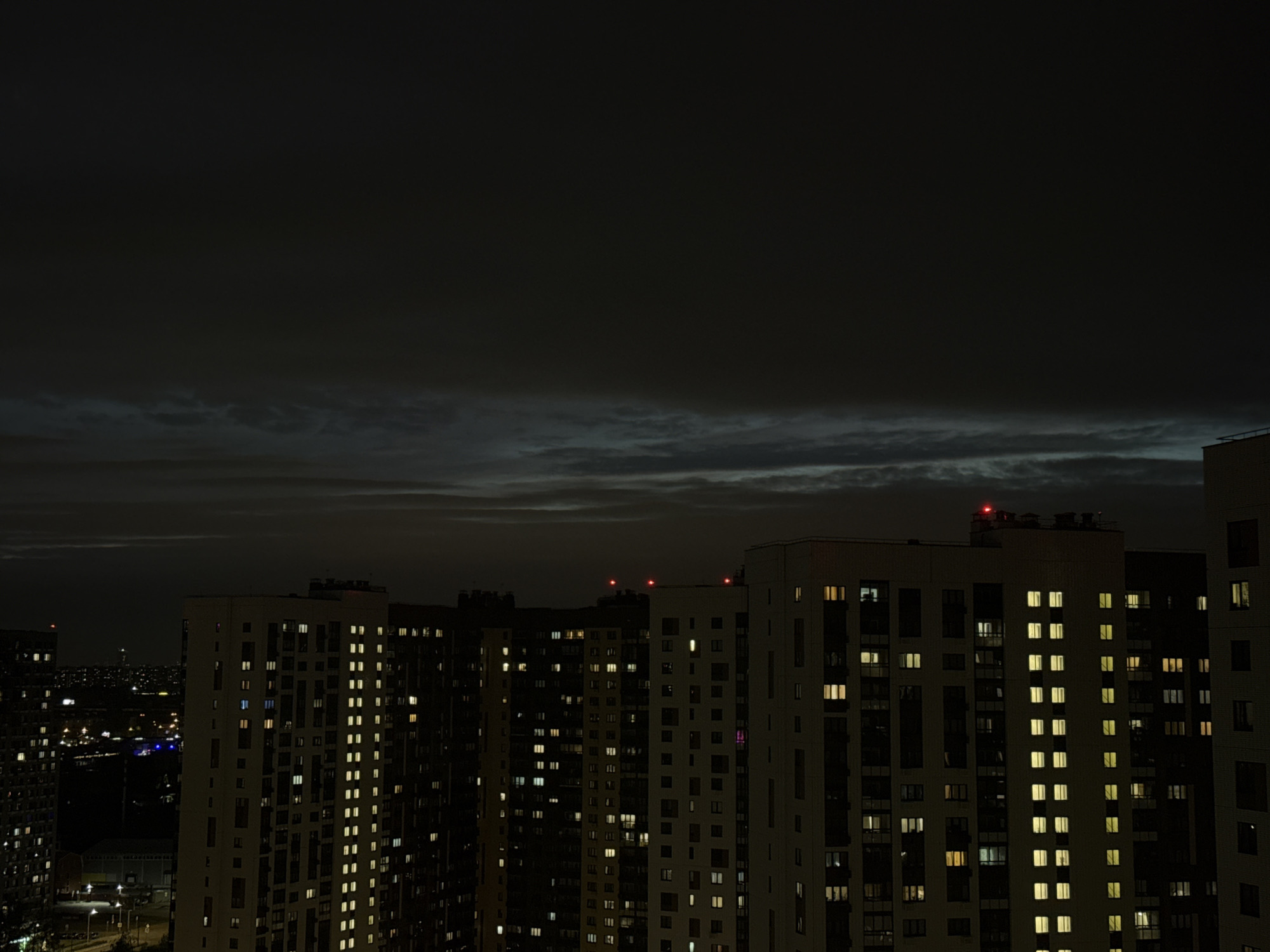 A night view shows densely packed high-rise apartment buildings. Many windows are illuminated with interior lights. Red lights are visible on the roofs of some buildings, likely navigation lights. In the background, there are scattered city lights. The sky is overcast. The photo was likely taken in Lyubertsy, Russia, given the appearance of the buildings and location tag. There are no people visible in the scene. The dominant elements are the architecture and urban landscape.