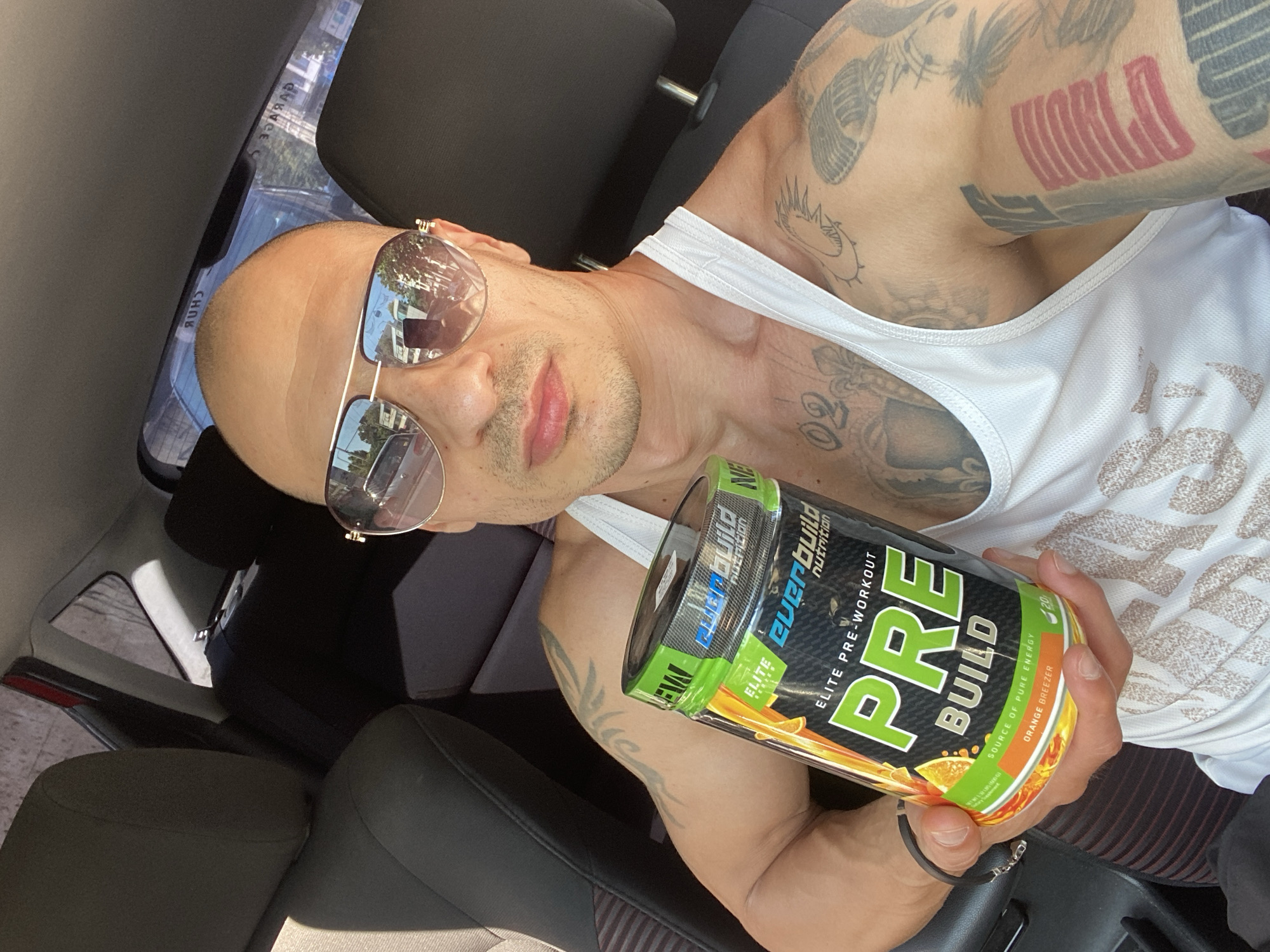 A man with a bald head, light stubble, and mirrored sunglasses is depicted in a close-up, selfie-style photograph taken inside a car. He is wearing a white tank top and his arms, shoulder, and neck are heavily tattooed with dark and red ink, including the word "WORLD" on his upper left arm. His expression is neutral, with his lips slightly parted.
He prominently holds a black and green "Everbuild Nutrition PRE BUILD Elite Pre-Workout" container towards the camera. The product label features text such as "SOURCE OF PURE ENERGY," "ORANGE BREEZER" (flavor), and "20" (likely servings), along with a "NEW" badge.
The setting is the interior of a vehicle, evident from the dark car seats and the angle of the shot. Bright sunlight indicates a clear, sunny day, with reflections in his sunglasses showing an outdoor urban environment, including buildings, trees, and blue sky. Faint reversed text, such as "GARAGE," is visible in the reflections, suggesting nearby signage. The photo is set in Pleven, Bulgaria, and captures the man presenting the supplement.
