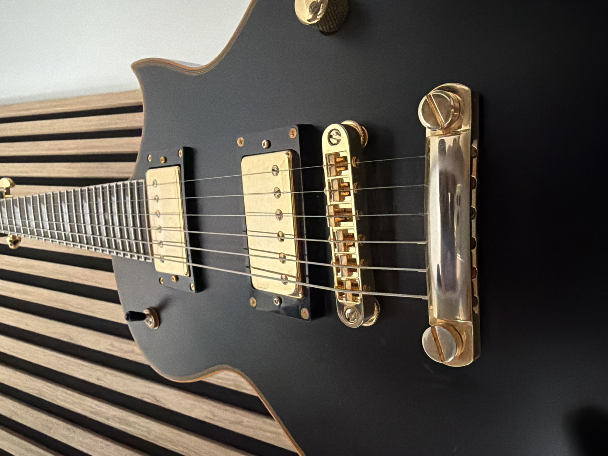 The image shows a close-up, side-view of a black electric guitar. The guitar has gold hardware, including the bridge, pickups, and tuning pegs. The fretboard is visible, with silver frets and white strings. The guitar is leaning against a wooden slatted wall. The lighting suggests it's daytime, likely indoors with artificial lighting. The scene is static, with no activity taking place beyond the guitar being displayed. No text is visible in the image. The context provided places the location in Łódź, Poland.