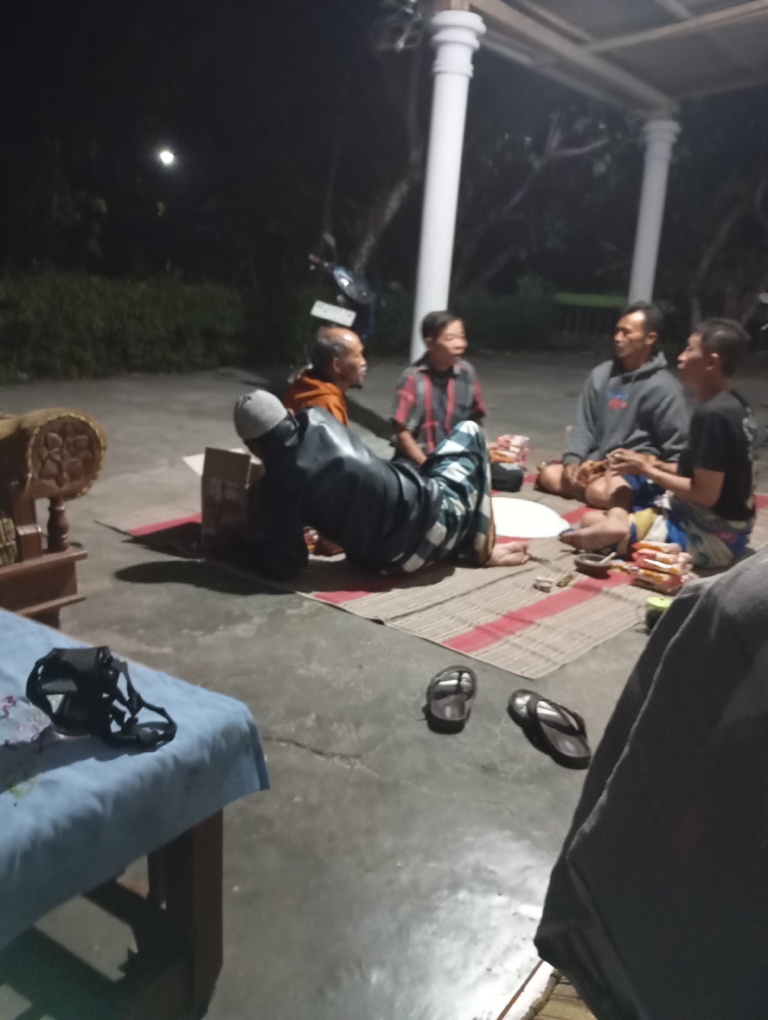 A group of four men are gathered outdoors at night, possibly on a patio or in a courtyard in Wonogondo, Indonesia. They are sitting on a woven mat on a concrete floor. The man closest to the foreground is lying on his back with his head propped up by a black object. He is wearing a white cap, a dark long-sleeved garment, and patterned pants. To his right, a man in an orange shirt is sitting upright, facing away from the camera. Next to him, another man wearing a plaid shirt and jeans is seated and appears to be talking. The fourth man is seated to his right, wearing a gray hoodie and jeans. There are several packages of what appear to be food or snacks on the mat in front of the seated men. In the background, there are trees and possibly a building with pillars. A table covered with a blue cloth is visible in the lower left corner of the image, with a black camera or strap resting on it. Two pairs of flip-flops are on the concrete floor in the foreground. The lighting suggests it is nighttime, with artificial lights illuminating the scene.