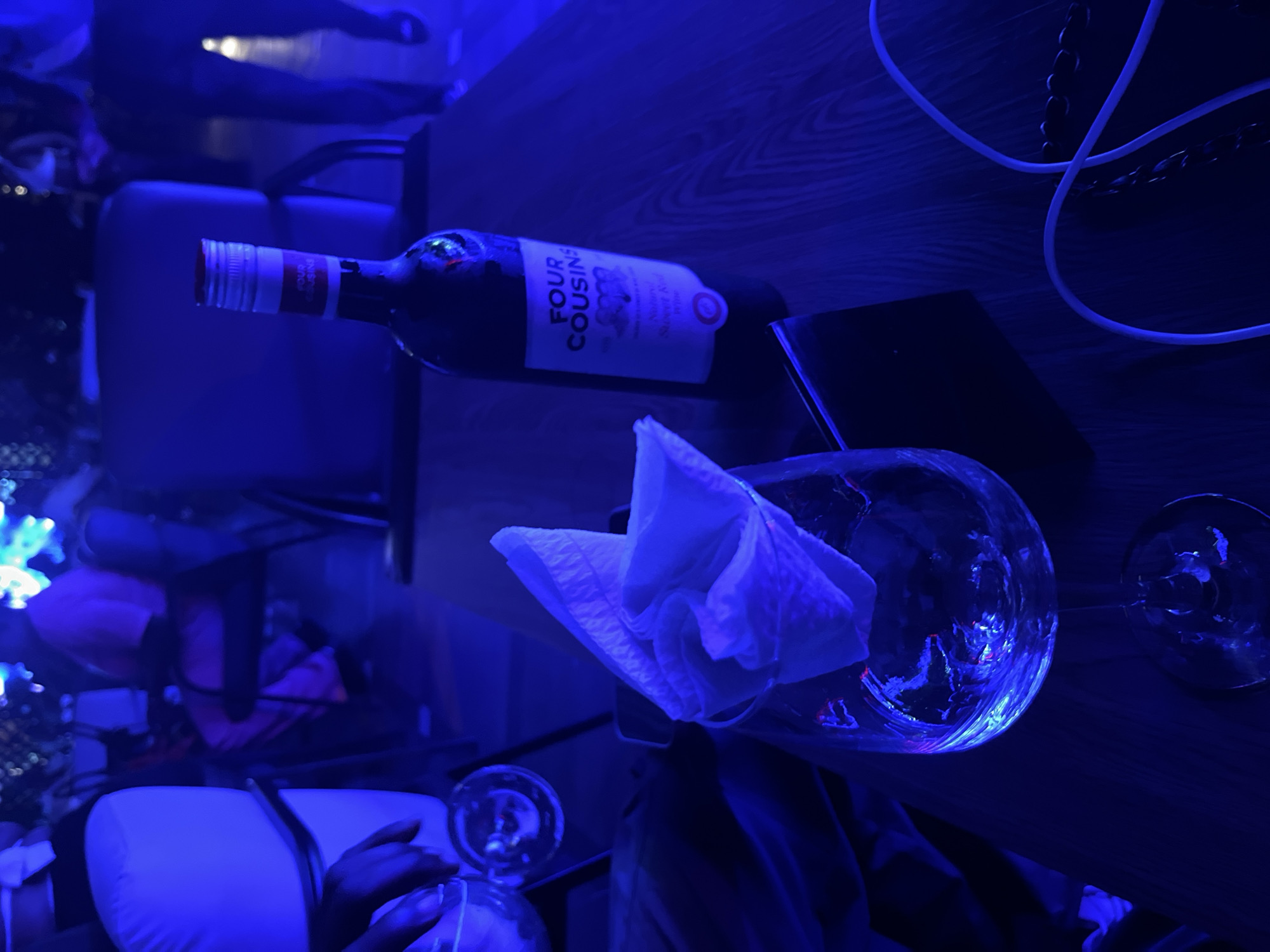 The image displays an indoor nighttime scene characterized by deep blue ambient lighting. In the foreground, a dark wooden table features a dark glass bottle of "FOUR COUSINS Natural Sweet Red Wine" with a white label and silver screw cap, appearing mostly full. Adjacent to it is an empty clear wine glass with a folded white paper napkin placed inside the bowl. To the left, a hand with dark skin tone holds a partially visible clear drinking glass. White electrical cables are visible on the right side of the table. The background contains blurred figures of multiple individuals; one person is seated in a dark chair wearing a light pink top, while another is partially visible walking on the far right. White padded seating and additional dark upholstered chairs are present in the background. The environment suggests a lounge or casual dining establishment. Specific identifiers for a precise location within Abuja, Nigeria, are not discernible from the visual evidence provided.