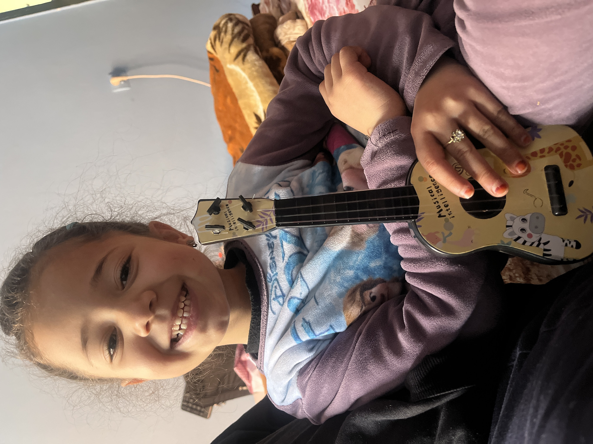 A young girl smiles directly at the camera while holding a toy ukulele with animal print. The background includes a light-colored wall, blankets, and other individuals partially visible. The location appears to be indoors, and there is no direct indication of being in Ouaouizert, Morocco.