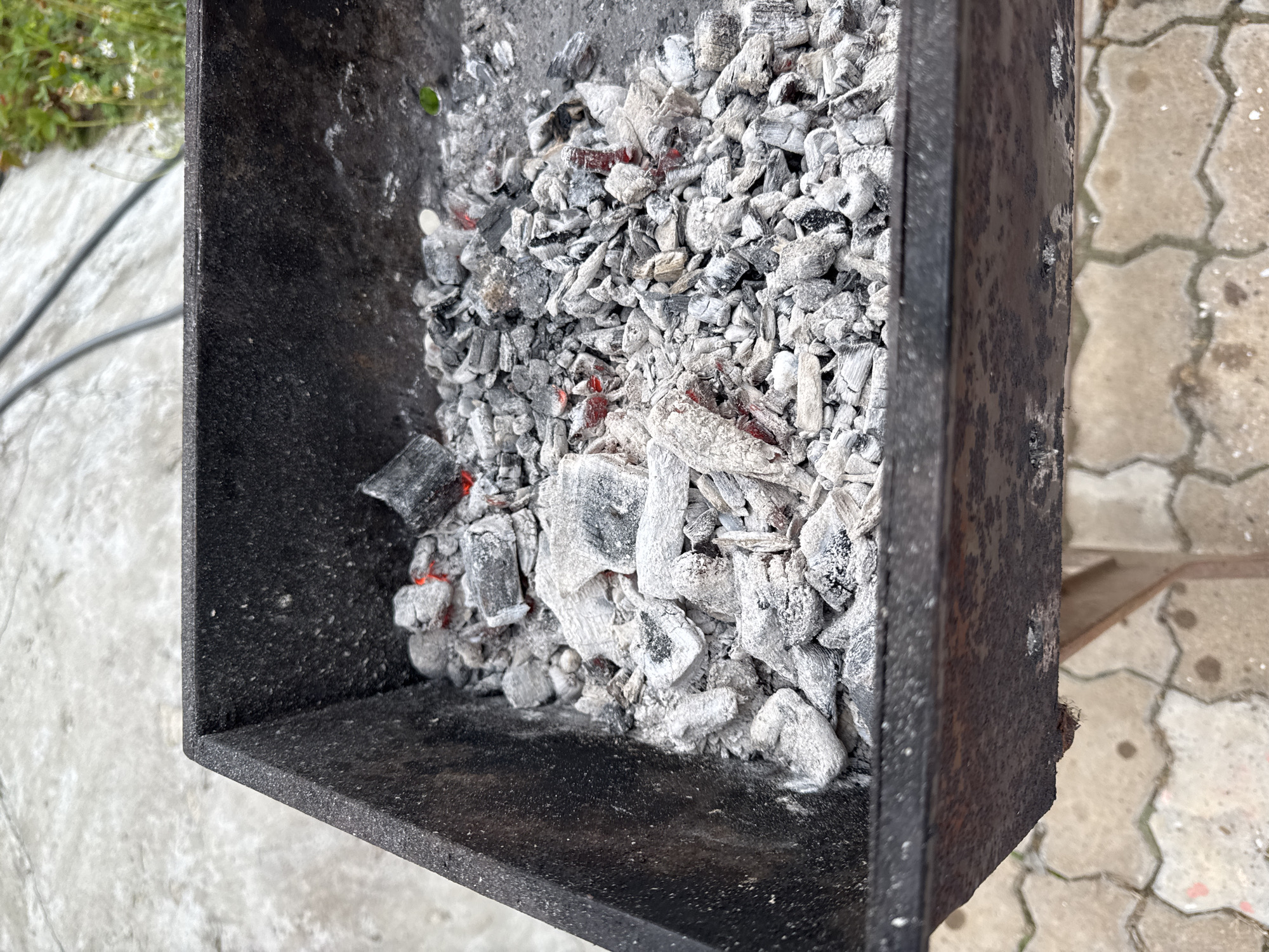 A close-up, overhead shot shows the inside of a dark metal barbecue grill filled with grey ash and charcoal remnants. Some pieces of charcoal still show glowing red embers. The grill is situated outdoors on a patterned concrete surface. The background includes more concrete and some out-of-focus green foliage. The location is identified as Chuguyevka, Russia.