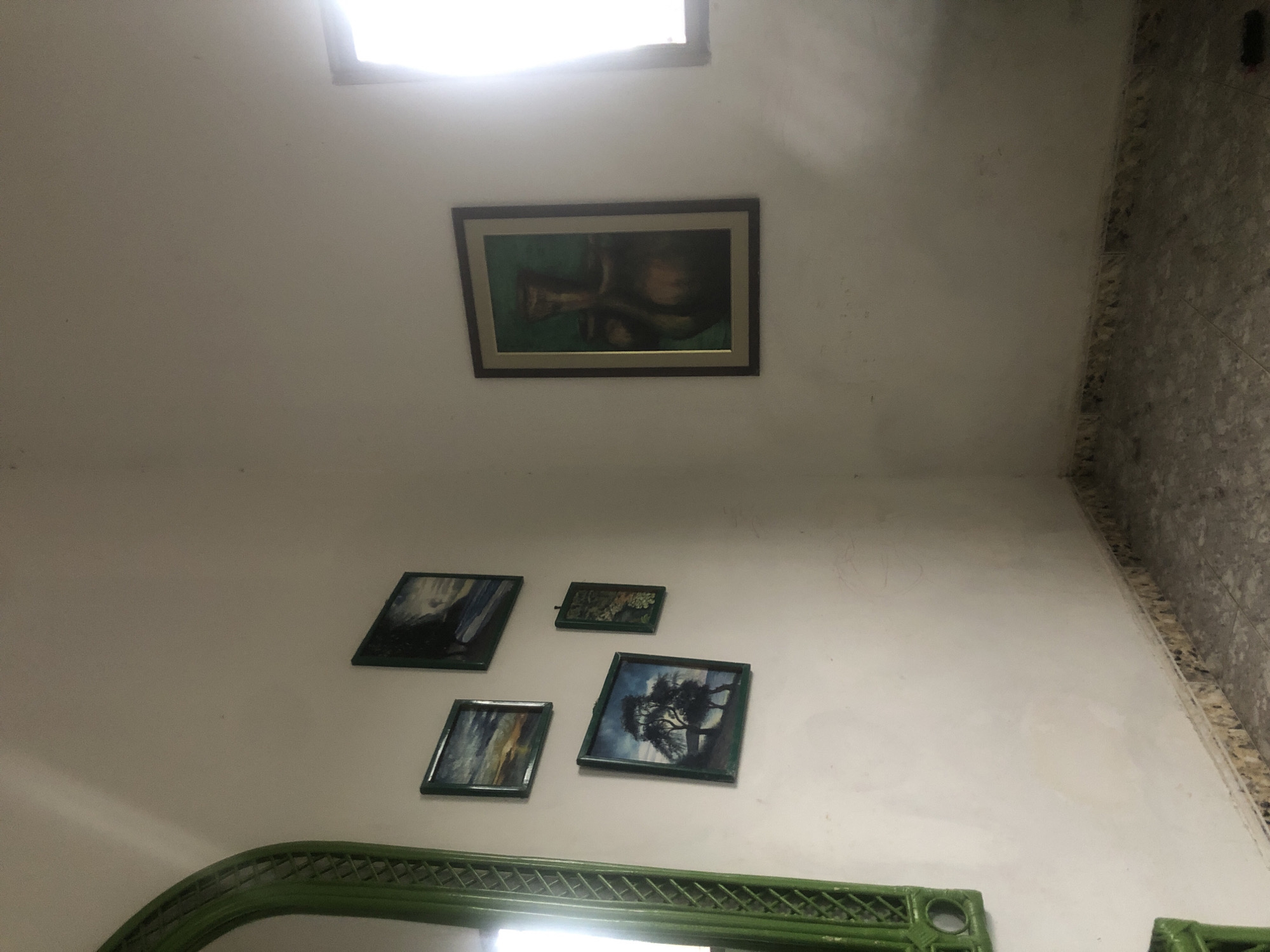 The image shows a corner of a room with white walls and a tiled floor. On the left wall, there is a large, ornate green mirror. To the right of the mirror, there are four framed paintings hanging on the wall. The paintings appear to be landscapes and abstract art. In the center of the room, on the right wall, there is a larger framed painting depicting a vase. To the right of this painting, there is a window with bright light streaming in. The overall impression is of a simple, decorated interior space. The location is identified as La Tagua, Venezuela.