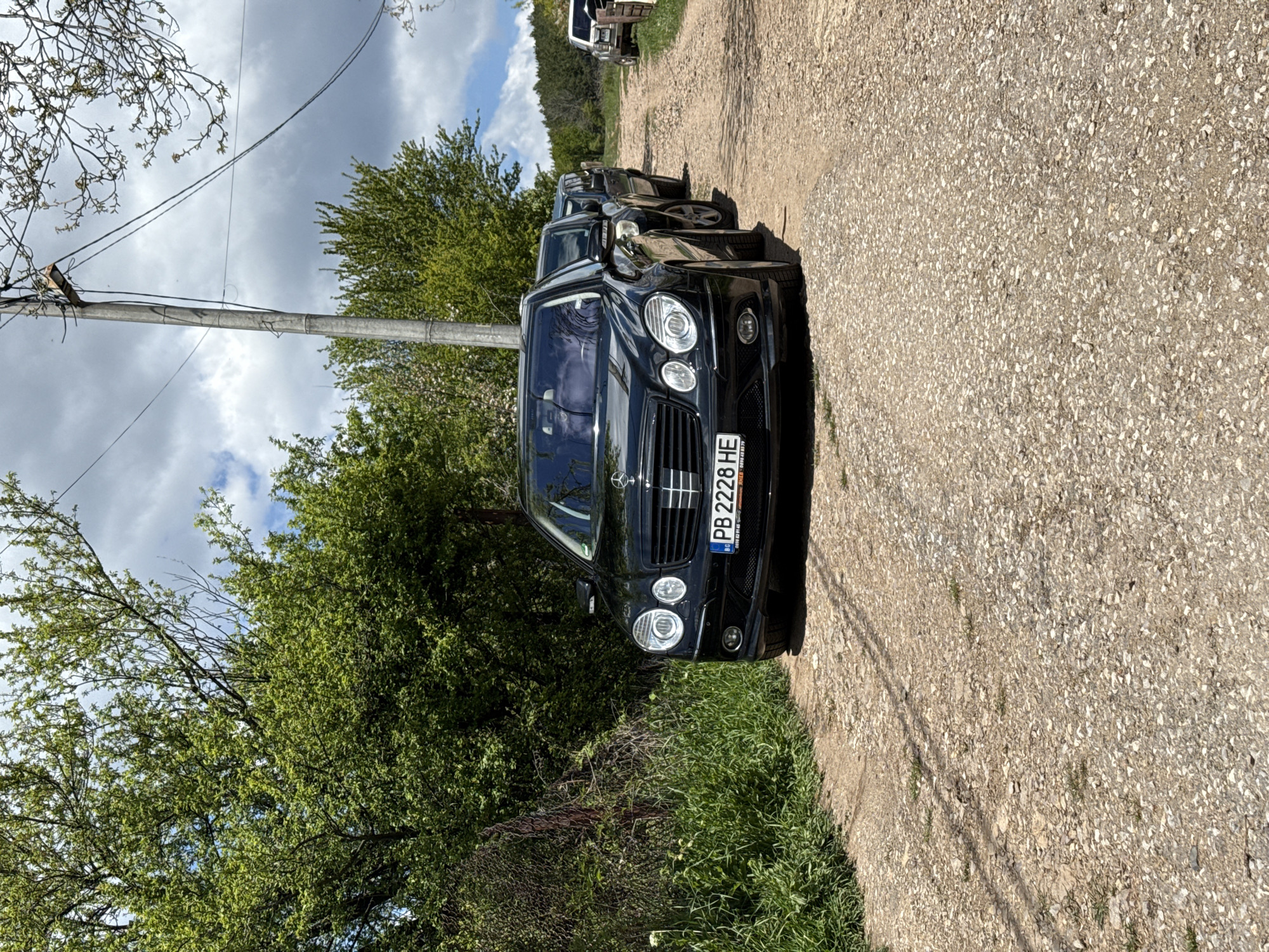 The image depicts a black Mercedes-Benz E-Class sedan parked on a dirt and gravel road in a rural or semi-rural setting near Buhovo, Bulgaria. Other vehicles are visible parked behind it. The car has a Bulgarian license plate reading "PB 2228 HE". The car is parked near a grassy area and a line of trees and foliage. A utility pole with wires extends into the scene, situated between the road and the greenery. The sky above shows a mix of clouds and patches of blue.