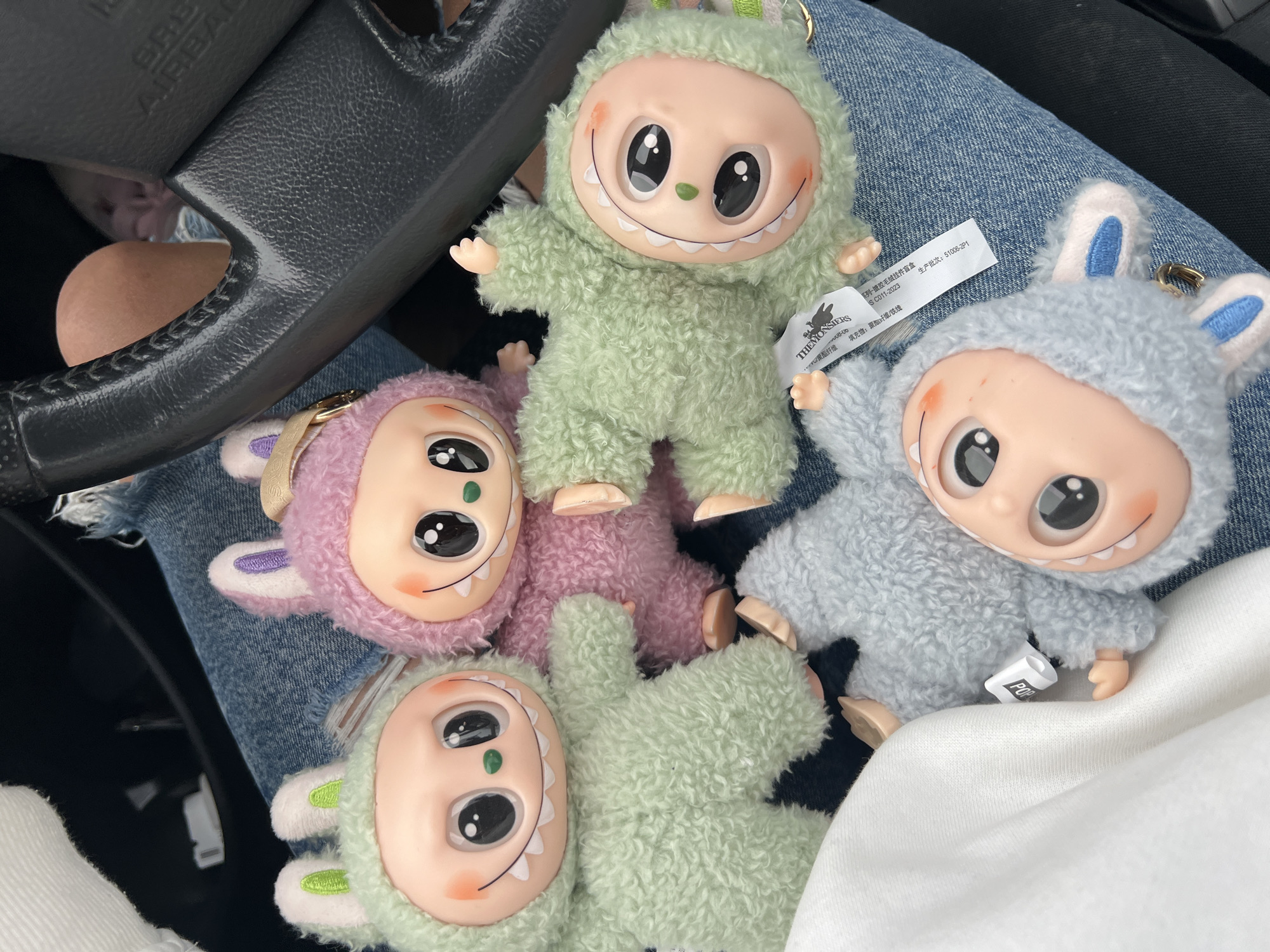 The image depicts four plush toy keychains resting on a person's lap inside a vehicle. The toys are stylized characters featuring large, light-skinned heads with wide smiles, prominent white teeth, large black eyes with white reflections, and small green noses. Each character is clad in a fuzzy, textured suit with ear-like appendages.
Specifically:
- One pale green toy with green-lined ears is positioned upright on the left.
- One pink toy with purple-lined ears and a small gold charm on its head is to the right of the first green toy.
- Another pale green toy with green-lined ears lies horizontally below the pink toy, with a white tag attached that reads "THE MONSTERS" in English and includes Chinese text.
- One light blue toy with blue-lined ears is positioned upside down on the lower left.
The toys are situated on a pair of light blue denim jeans, which show visible fraying along a seam in the upper left. A white fabric, possibly a garment, is visible on the far left. A dark gray steering wheel, featuring visible stitching and an "AIRBAG" label, is partially visible on the right side of the frame, indicating the setting is a car interior. The upper thigh or knee of an individual is partially visible behind the steering wheel.
The image does not contain any visual information to determine a specific location within the city of Budva, Montenegro.