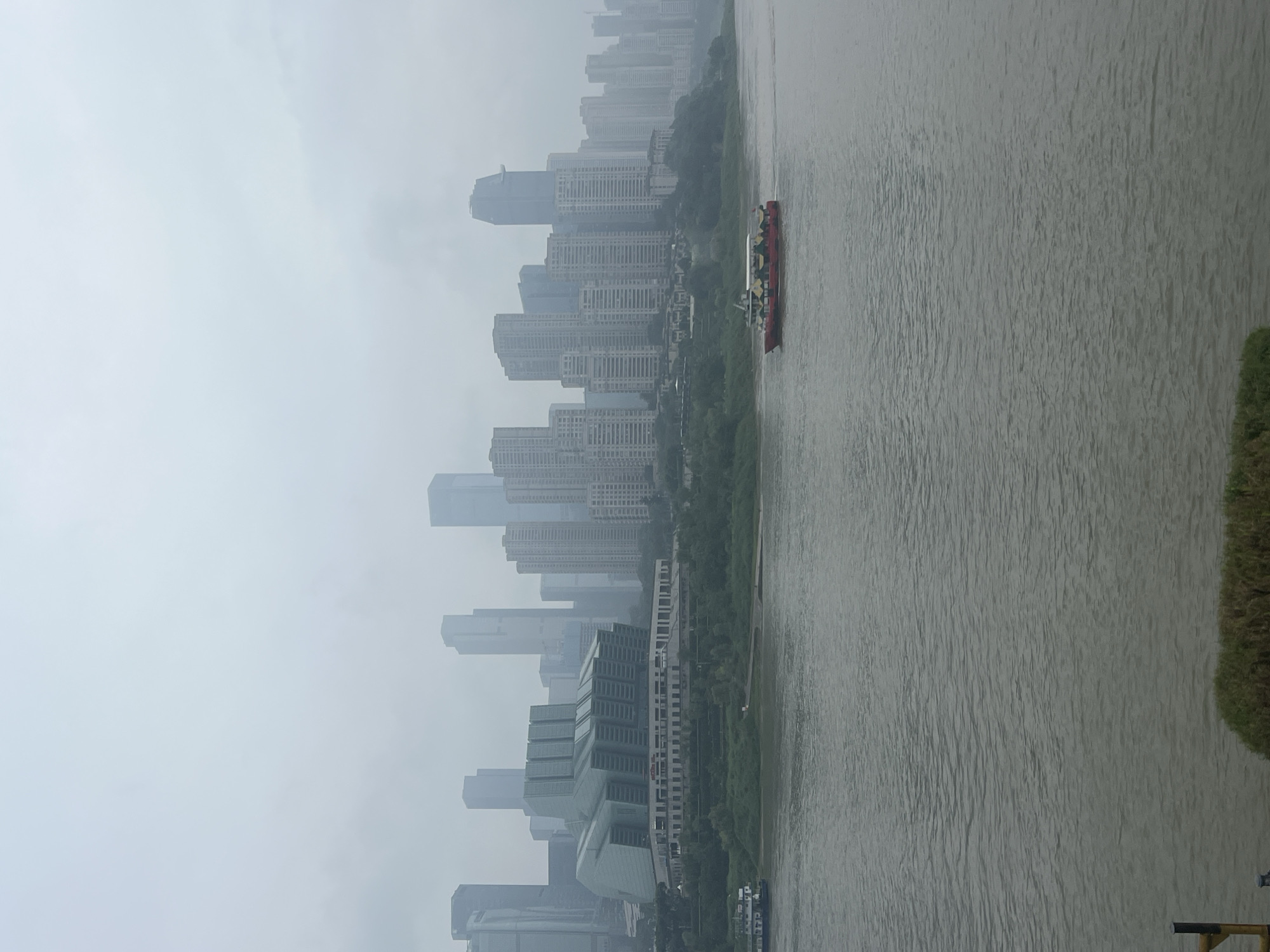 The image depicts a cloudy, hazy day overlooking a wide river and a dense cityscape. On the river, two boats are visible: a white boat on the left side and a red and yellow boat further to the right, moving towards the right edge of the frame. The cityscape, characteristic of Yuzhong District, China, features numerous tall buildings, including skyscrapers and a distinctive green-domed building in the foreground. Vegetation lines the riverbank between the water and the buildings. The overall atmosphere is subdued due to the overcast sky and atmospheric haze, obscuring details of the distant buildings.