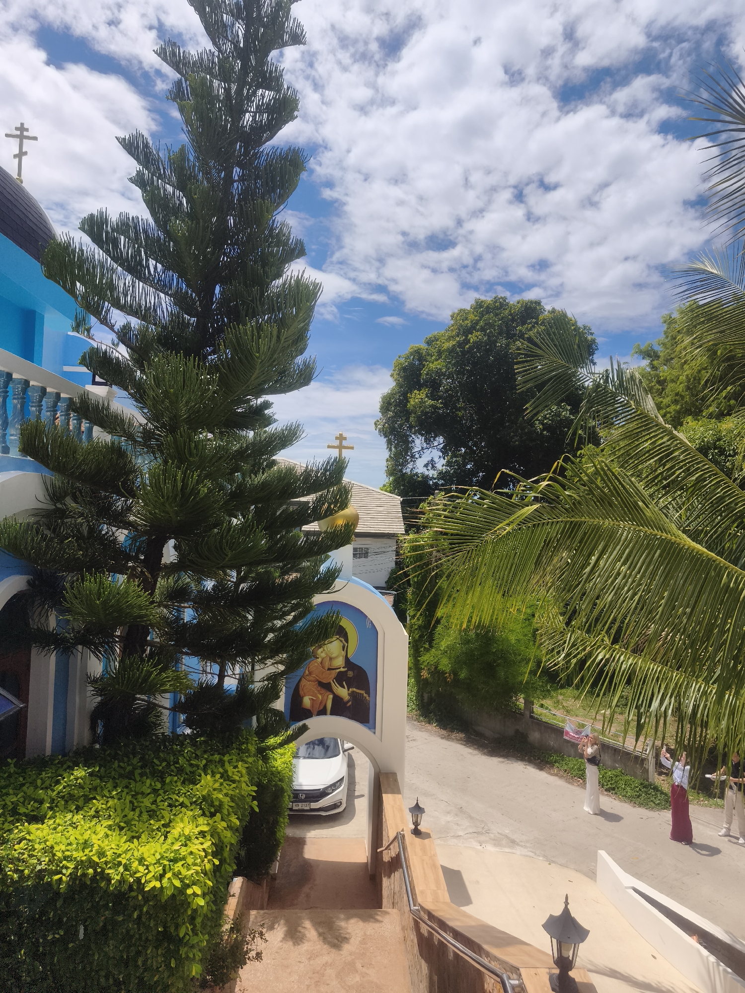 The image depicts an outdoor scene in Ko Samui, Thailand, captured from an elevated perspective, likely the steps or a balcony of a building. The sky is bright blue with scattered white cumulus clouds.
On the left side, a prominent blue and white structure, consistent with an Orthodox church, is visible. It features a dark domed roof topped with a silver Orthodox cross. A second, smaller golden Orthodox cross is visible on a white roof further back, behind a large conifer tree. The church's facade includes white balustrades and an arched entrance with a painted icon of the Virgin Mary holding the infant Christ. At the base of the church, manicured green bushes line a pathway where a white Honda Civic sedan (license plate "4B 2137" partially visible) is parked beneath an archway.
A tall, dense conifer tree stands centrally in the mid-ground, partially obscuring the church. Lush tropical vegetation, including large dark green trees and prominent bright green palm fronds, fills the right side and background of the image.
In the lower right and mid-ground, a light-colored asphalt or concrete road curves through the scene. Several individuals are present on this road: one woman in a white dress with a dark top is holding a framed object or banner, apparently posing for a photograph. To her right, another woman wearing a light blue top and a long maroon skirt is visible, also seemingly posing, with a man in a dark shirt and light pants standing further right, holding a device. A low, white fence runs along the roadside.
The foreground on the right features terracotta-colored steps with a metal handrail and decorative outdoor lamps with dark metal bases and frosted glass shades. The scene is brightly illuminated by sunlight, indicating clear weather.