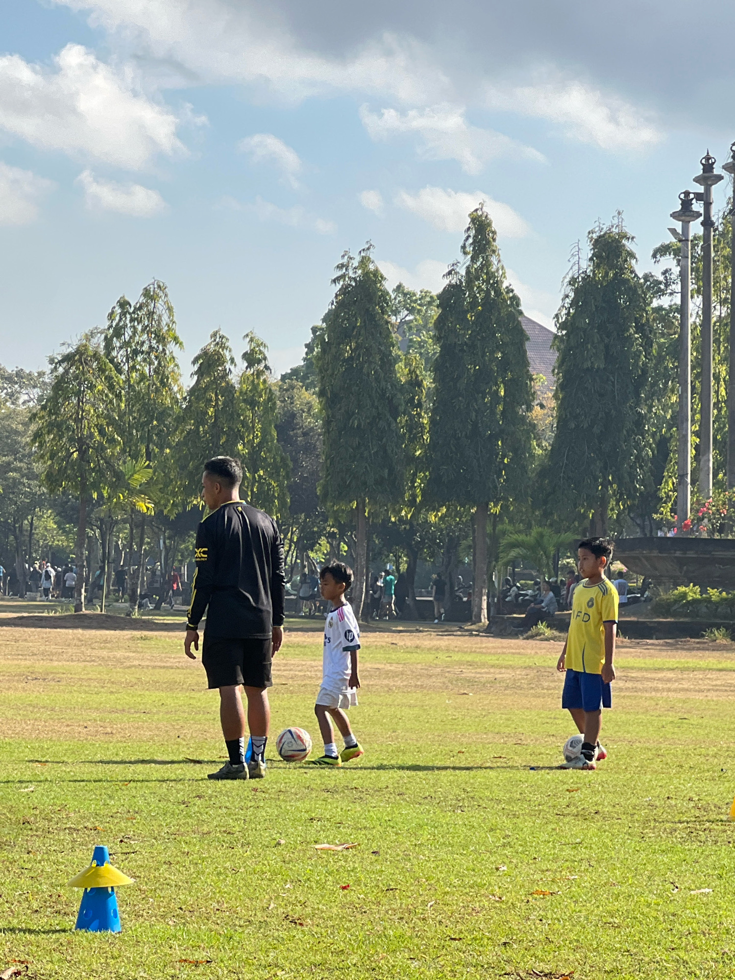 This image depicts a sunny outdoor scene on a grassy field, likely in Denpasar, Indonesia, featuring a group of people engaged in what appears to be a soccer training session. In the foreground, a man wearing a black long-sleeved shirt with yellow accents and black shorts stands facing left, appearing to observe or instruct. To his right, a young boy in a white soccer jersey with blue shorts and a red soccer ball at his feet is also present. Further to the right, another young boy in a yellow soccer jersey with blue shorts stands near another soccer ball. The background is dominated by a line of tall, slender trees with dense green foliage, characteristic of tropical vegetation. Beyond the trees, there are hints of buildings with tiled roofs and possibly some decorative structures or statues, suggesting a park or a recreational area within the city. The sky is a light blue with scattered white clouds, indicating fair weather. Several other individuals are visible in the mid-ground and background, suggesting a larger gathering or multiple activities taking place on the field. A blue and yellow cone-shaped marker is positioned in the lower left corner of the frame, further indicating a training or practice setting. The overall impression is one of a vibrant community space being utilized for sports and recreation under a clear sky.