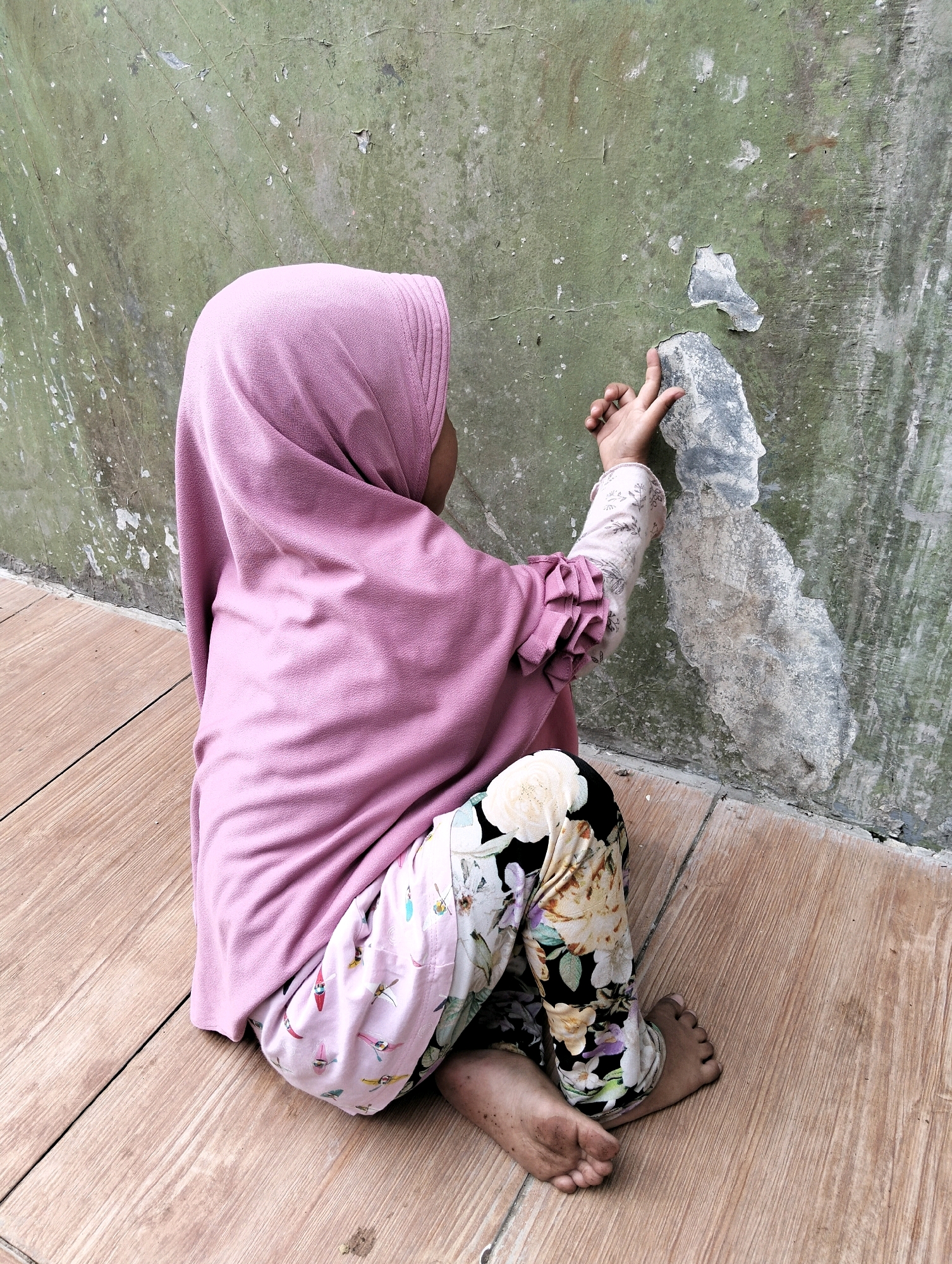 A young child wearing a pink hijab, a long-sleeved shirt with ruffles at the wrists, and patterned pants sits on a wooden floor next to a distressed green wall. The child's right hand is raised, with fingers touching a patch of peeling paint on the wall. The wall shows signs of age and wear, with cracked and discolored sections. The wooden floor is made of planks laid side by side. The overall scene suggests an outdoor or semi-outdoor environment.