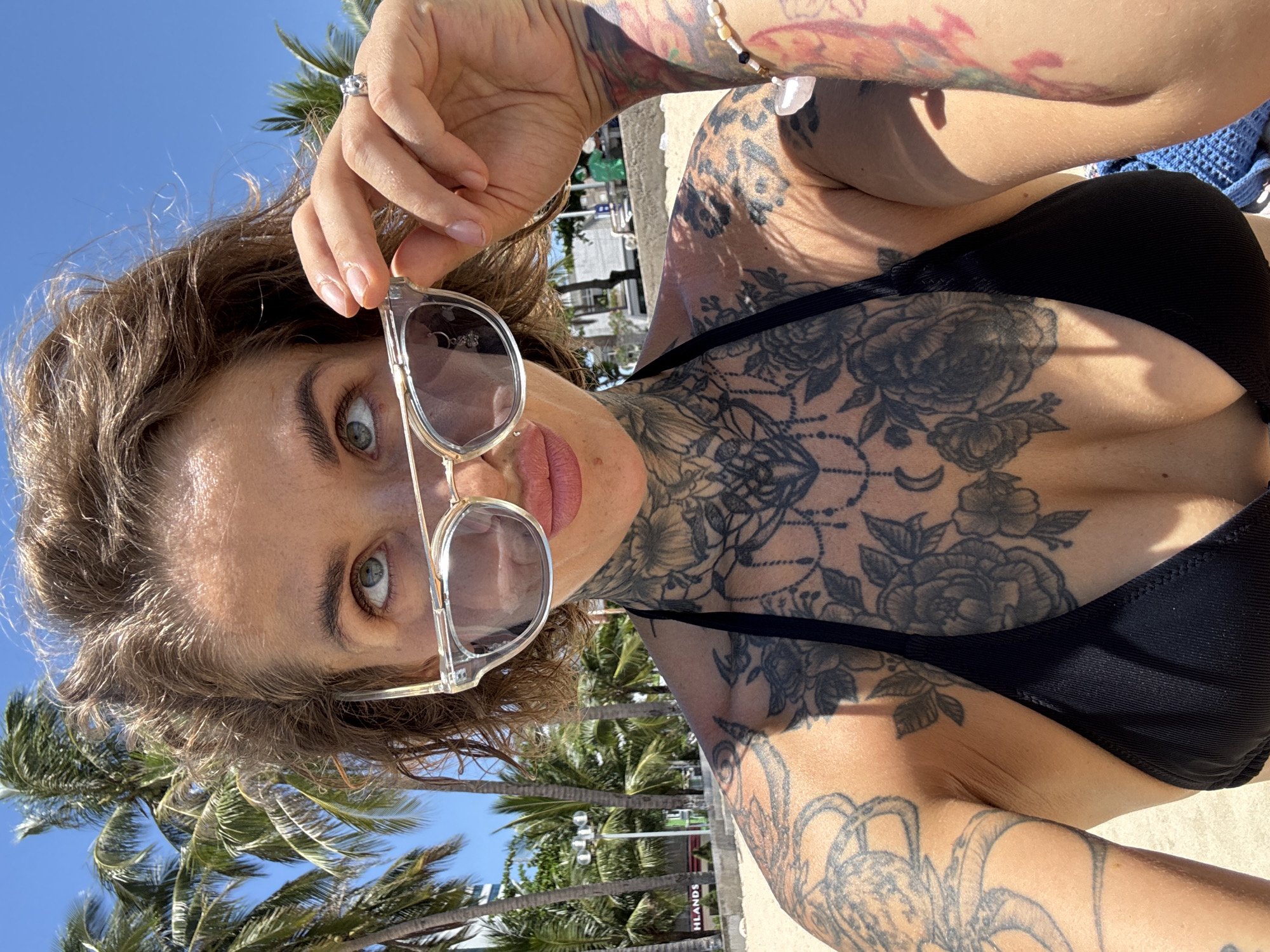 This is a medium-shot, selfie-style photograph taken outdoors on a sunny day. The subject is a woman with short, tousled brown hair, her face angled upwards and to her left, with her eyes looking in the same direction. She is wearing clear-rimmed sunglasses, which she is holding up with her right hand. She has a black bikini top on, and extensive floral and other tattoo work is visible on her chest and arms. The background shows a sandy beach with palm trees and buildings, consistent with a coastal urban environment. The location is identified as Bac Nha Trang Ward, Vietnam.