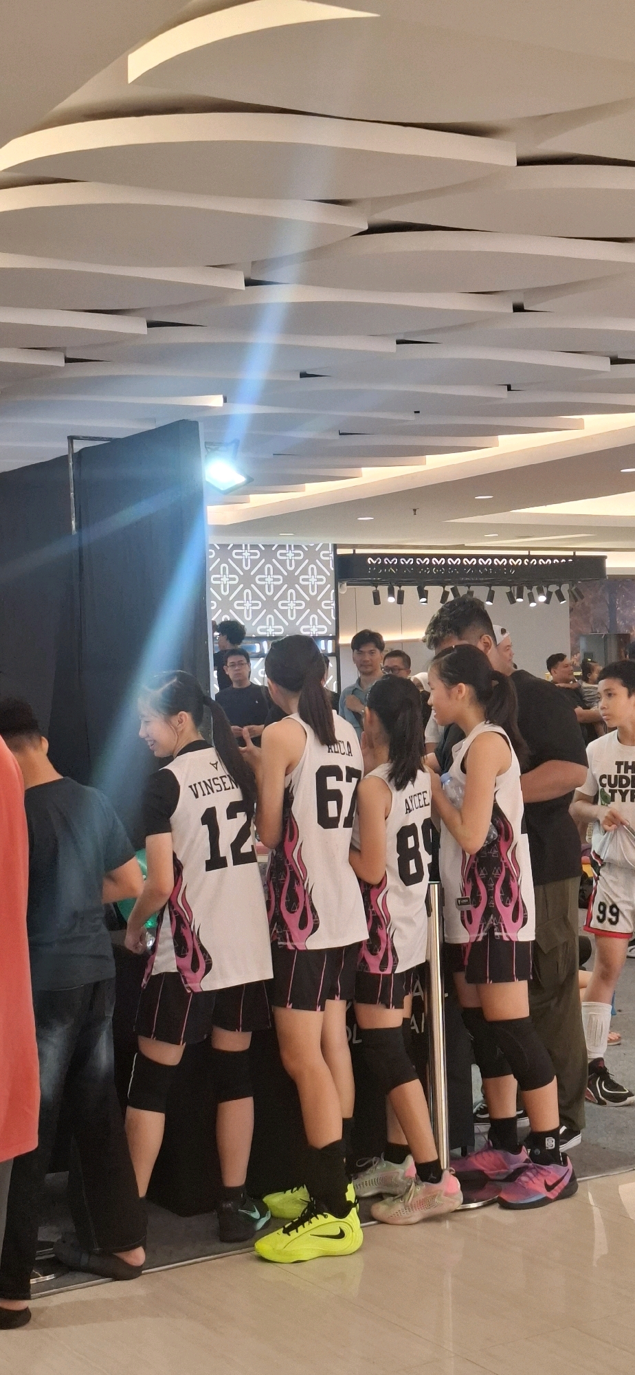 The image captures a group of young women, likely athletes based on their attire, standing in what appears to be an indoor public space, possibly a mall in Grogol, Indonesia, given the context. The women are positioned around a black curtain or booth, with some facing away from the camera and others turned slightly towards it. They are wearing matching white sports jerseys with pink flame designs and black shorts with pink trim. The jerseys display numbers and names, with visible numbers including "12," "67," and "89." Some of the women are wearing knee pads. In the background, other individuals, presumably spectators or staff, are visible. The environment features a modern, illuminated ceiling with a distinctive wavy design. The lighting is bright, and a visible light streak crosses the scene. A patterned wall or backdrop is also present behind the group. The floor is reflective, suggesting a polished surface. The overall impression is of a public event or gathering.