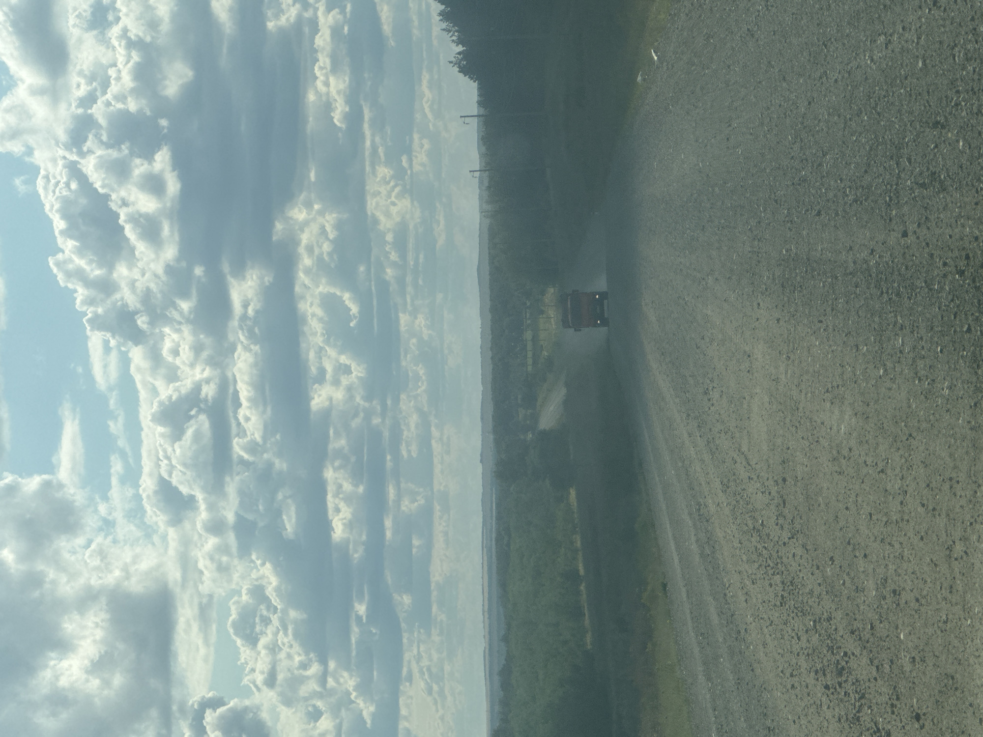 The image depicts a rural road in Vasilievka, Russia, with a truck driving away from the camera. The road is unpaved and covered in gravel, with a slight incline. The surrounding environment is characterized by dense vegetation, consisting of trees and shrubs, extending into the distance. The sky is partly cloudy, with a mix of white and gray clouds against a pale blue backdrop. The overall lighting suggests it is daytime, possibly late afternoon or early morning, given the soft light. There are no visible people besides the driver within the truck, and no other significant structures or objects present.