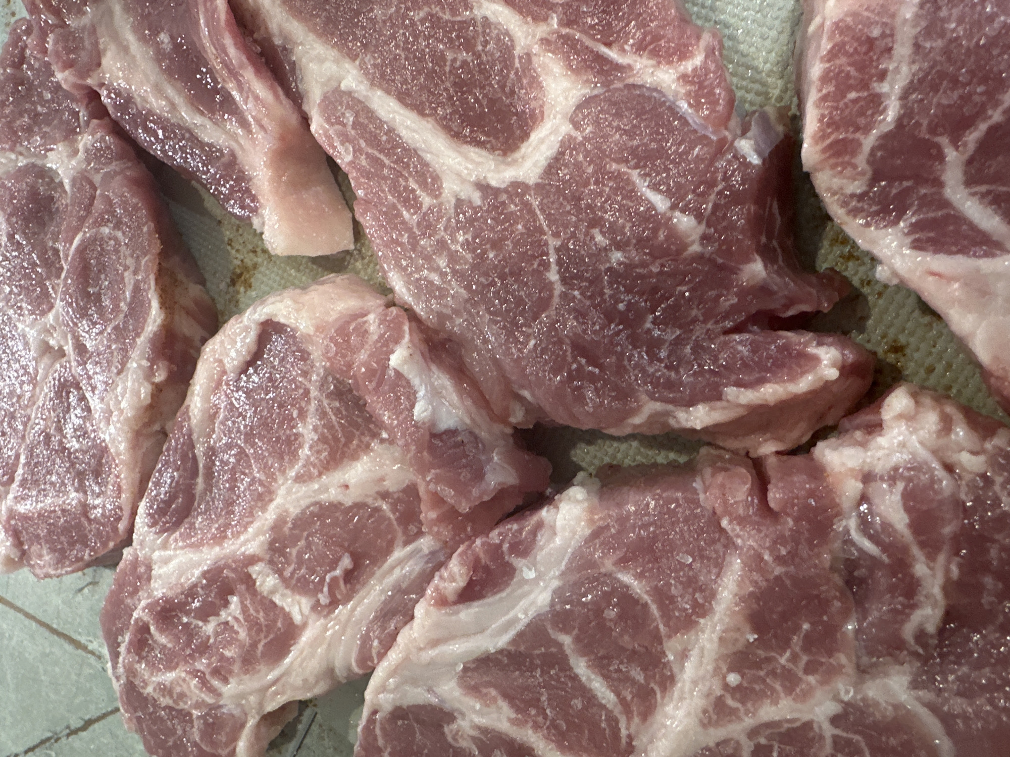 The image shows a close-up of several pieces of raw pork, likely pork shoulder or butt due to the marbling and fat content. The meat is predominantly pinkish-red with streaks of white fat running through it. The pieces are arranged somewhat haphazardly on a surface that appears to be a slightly textured, light-colored tray or cutting board. There are no people visible, nor is there any context to ascertain a city or location.