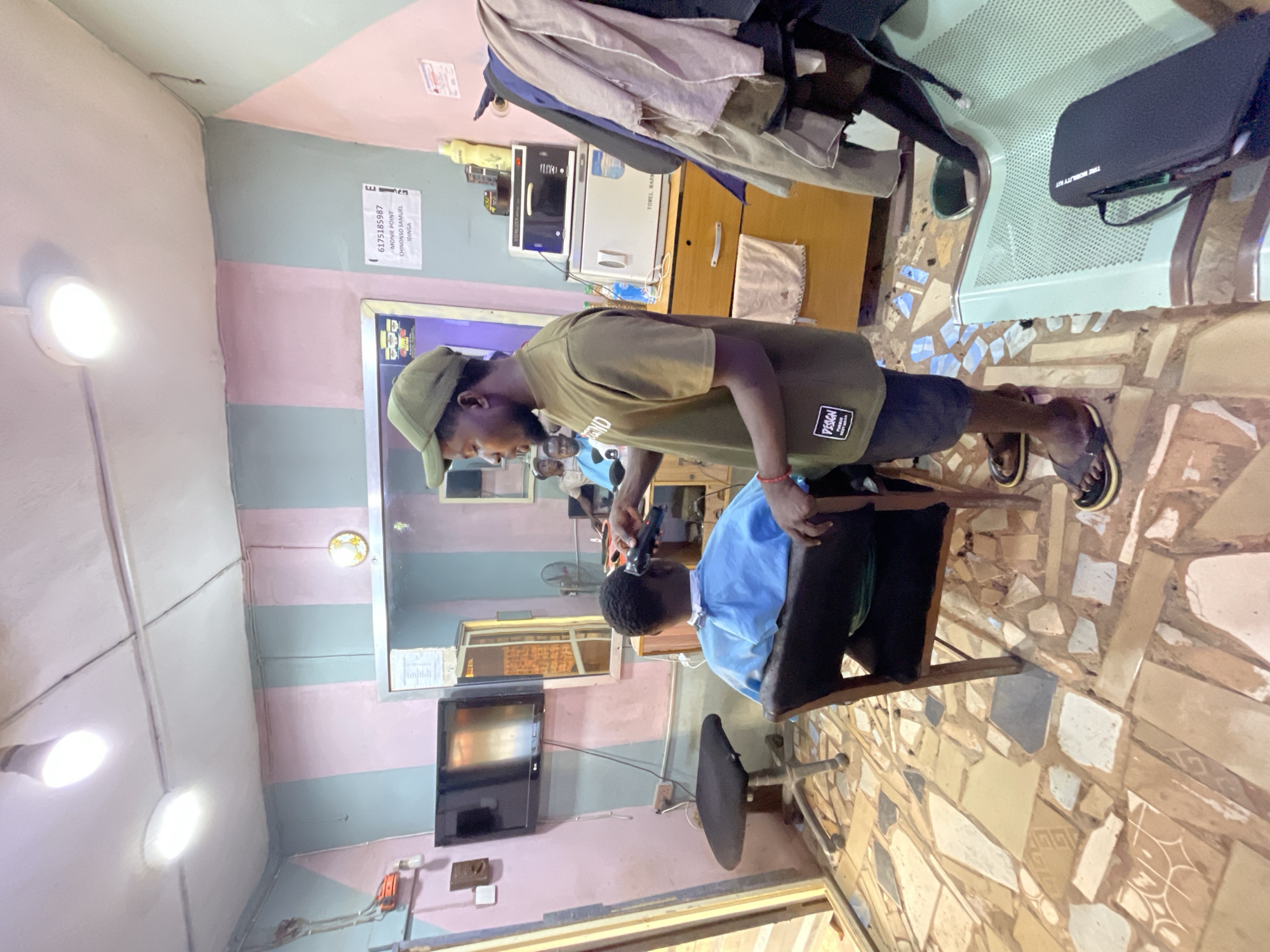 Inside a barbershop in Onitsha, Nigeria, a barber wearing a green cap and an olive-green shirt is giving a haircut to a young client seated in a black chair. The barber holds an electric hair clipper. The room's decor includes striped walls, a television on the wall, and a tiled floor. A sterilizer and other barber tools are on a shelf in the background. Clothes are hanging from a rack near the wall. A phone number and a name tag are visible on the wall.