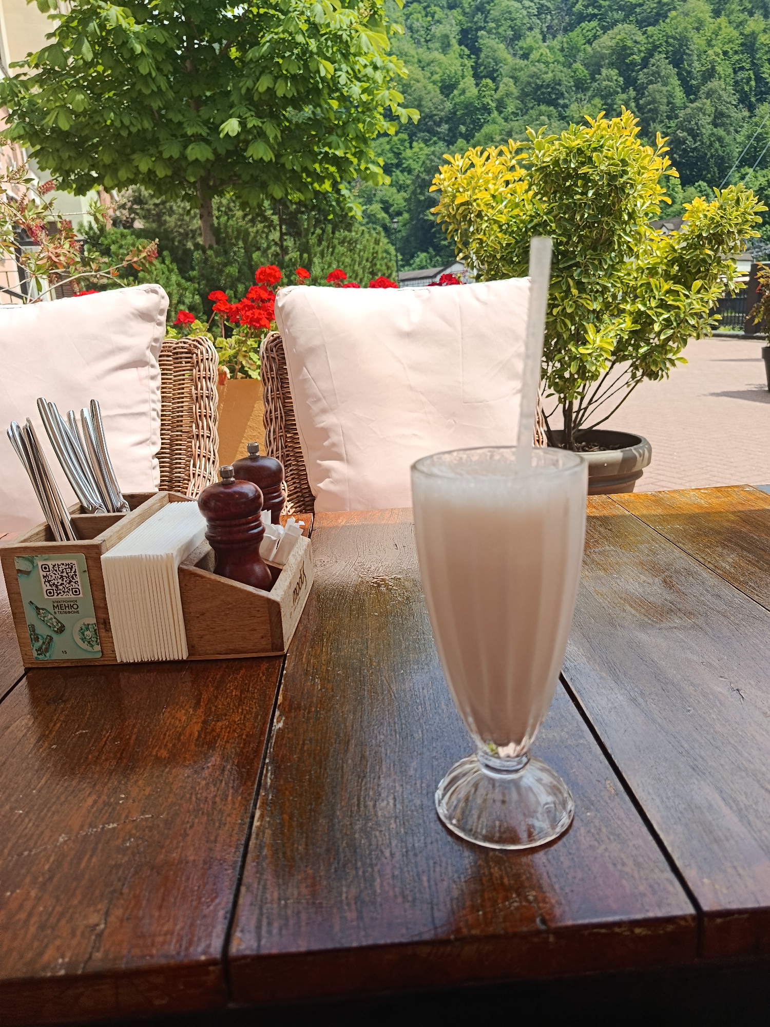 A close-up, eye-level, outdoor shot captures a wooden table in a sunlit restaurant setting in Estosadok, Russia. On the table, a tall glass filled with a pale, frothy beverage and a white straw is positioned on the right side. To the left of the drink is a wooden caddy containing silverware, a stack of white napkins, and a pair of dark wooden salt and pepper grinders. In the background, wicker chairs with light-colored cushions are visible, along with lush green trees and red flowers, suggesting a natural and pleasant atmosphere. The overall scene conveys a relaxed outdoor dining experience.