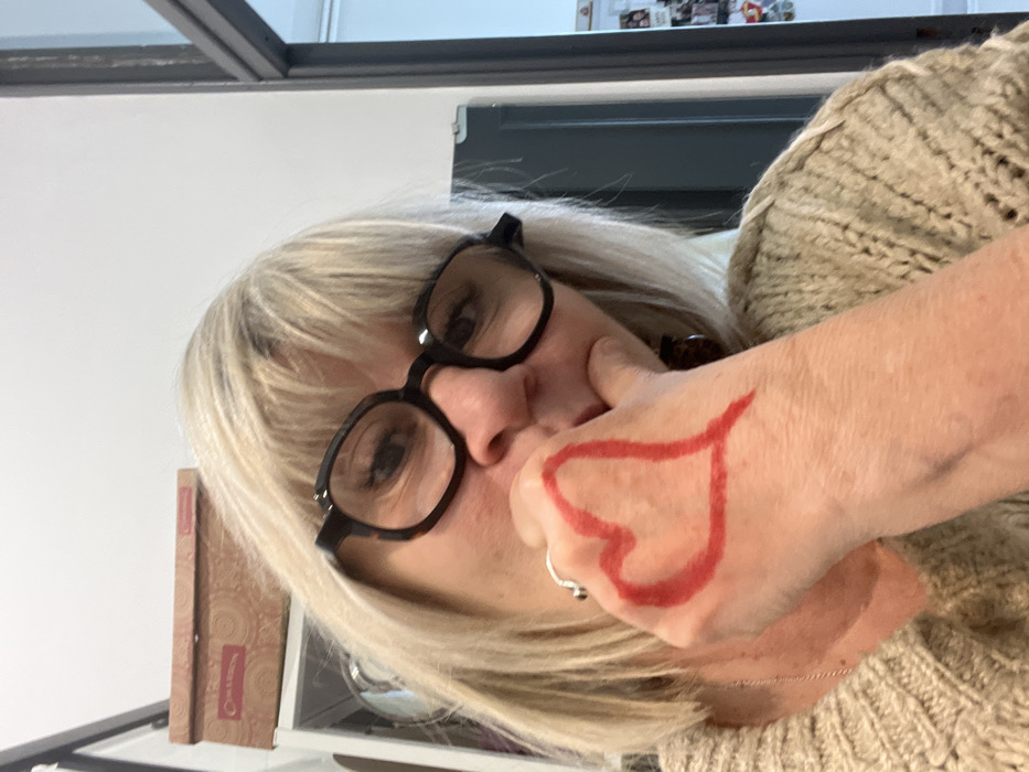 A blonde individual, appearing to be female, is depicted in a close-up, chest-up view, looking directly towards the viewer. She wears dark-rimmed glasses and a light-colored, possibly beige or oatmeal, chunky knit sweater. Her left hand is prominently displayed in the foreground, with a red heart shape drawn on the back of it. A silver-toned ring is visible on her left ring finger. Her left hand's knuckles are near her chin or lower jaw, suggesting a contemplative or casual pose. To the immediate left of her head in the background, a brown box with a patterned design and the word "Charmon" (or similar) is visible on a white shelf. The background consists of a plain white wall and, to the right, a dark-framed doorway or partition revealing a glimpse of another room, possibly with a collage of small items or photos on a far wall. The specific location within Montélimar, France, cannot be determined from the visual evidence provided.