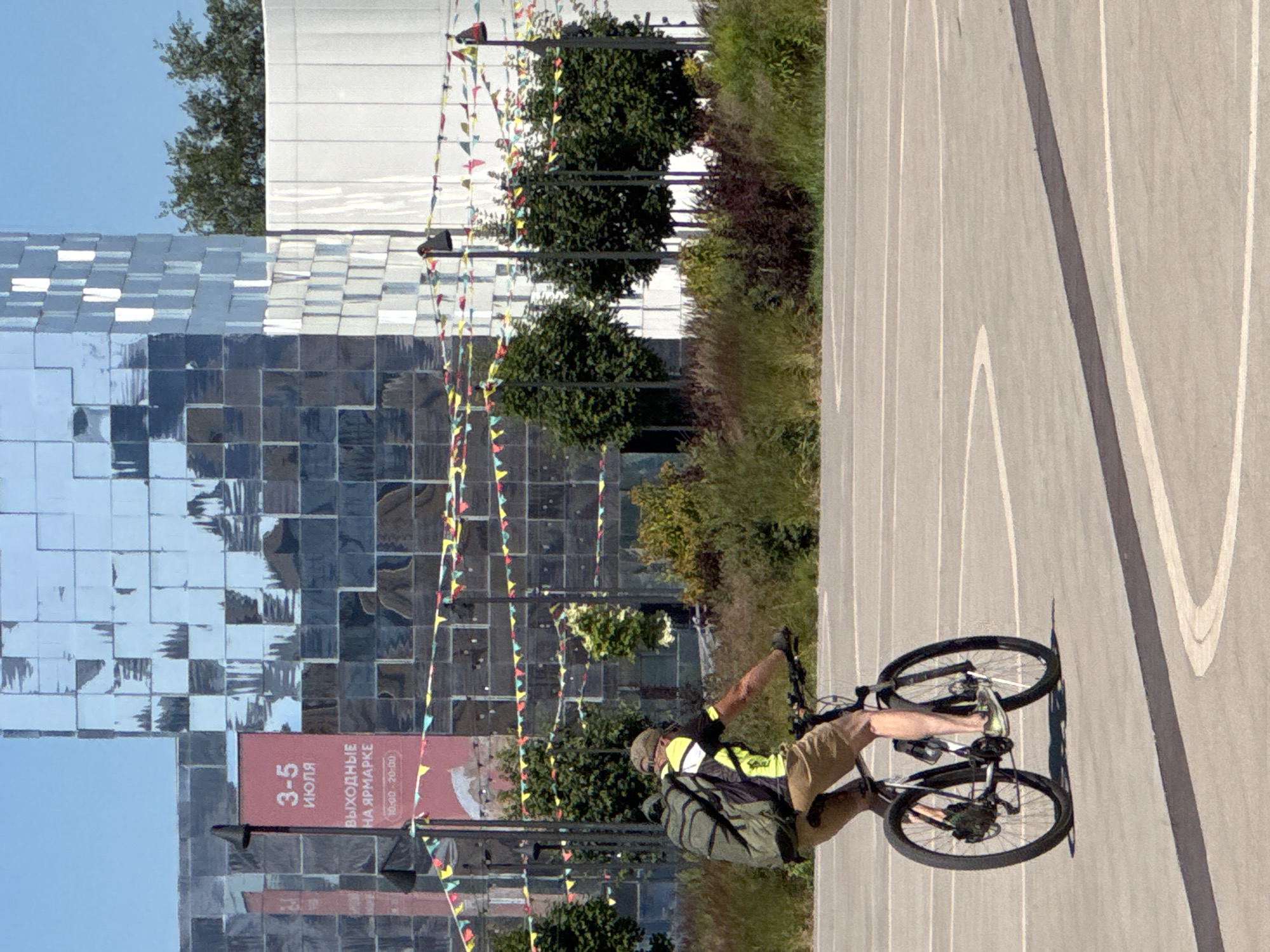 A person wearing a cap, t-shirt, and shorts rides a bicycle on a paved area with white markings. The person has a backpack on. In the background, a modern building with a mirrored facade stands under a clear blue sky. Decorative flags are strung between poles in front of the building. The scene appears to be outdoors during daylight. A banner on a pole near the building displays Cyrillic text, including "3-5 ИЮЛЯ" (July 3-5) and "ВЫХОДНЫЕ НА ЯРМАРКЕ" (Weekend at the fair), suggesting the location is in Nizhny Novgorod, Russia.