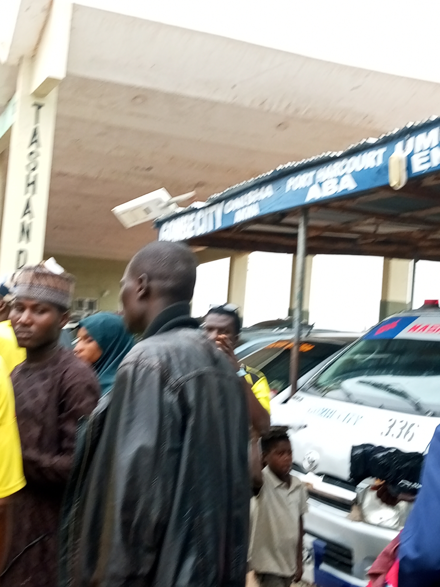 In Gombe, Nigeria, a scene unfolds under a covered walkway where several individuals are gathered around a vehicle. A prominent blue sign above reads "GOMBE CITY - ONITSHA - PORT HARCOURT - ABA". To the left, a pillar has the word "TASHA" vertically inscribed on it. In the foreground, three men are visible. The man on the left wears a patterned brown garment and a cap. Next to him, a woman in a blue hijab is partially obscured. A third man, facing away from the camera, is wearing a black leather jacket. Behind these individuals, a white vehicle, possibly an ambulance, is parked. The vehicle has red and blue markings and the number "336" is visible on its side. A young boy in a light brown shirt stands near the vehicle. The environment appears to be an urban setting, likely a public space given the signage and the presence of a vehicle. The lighting suggests it is daytime.