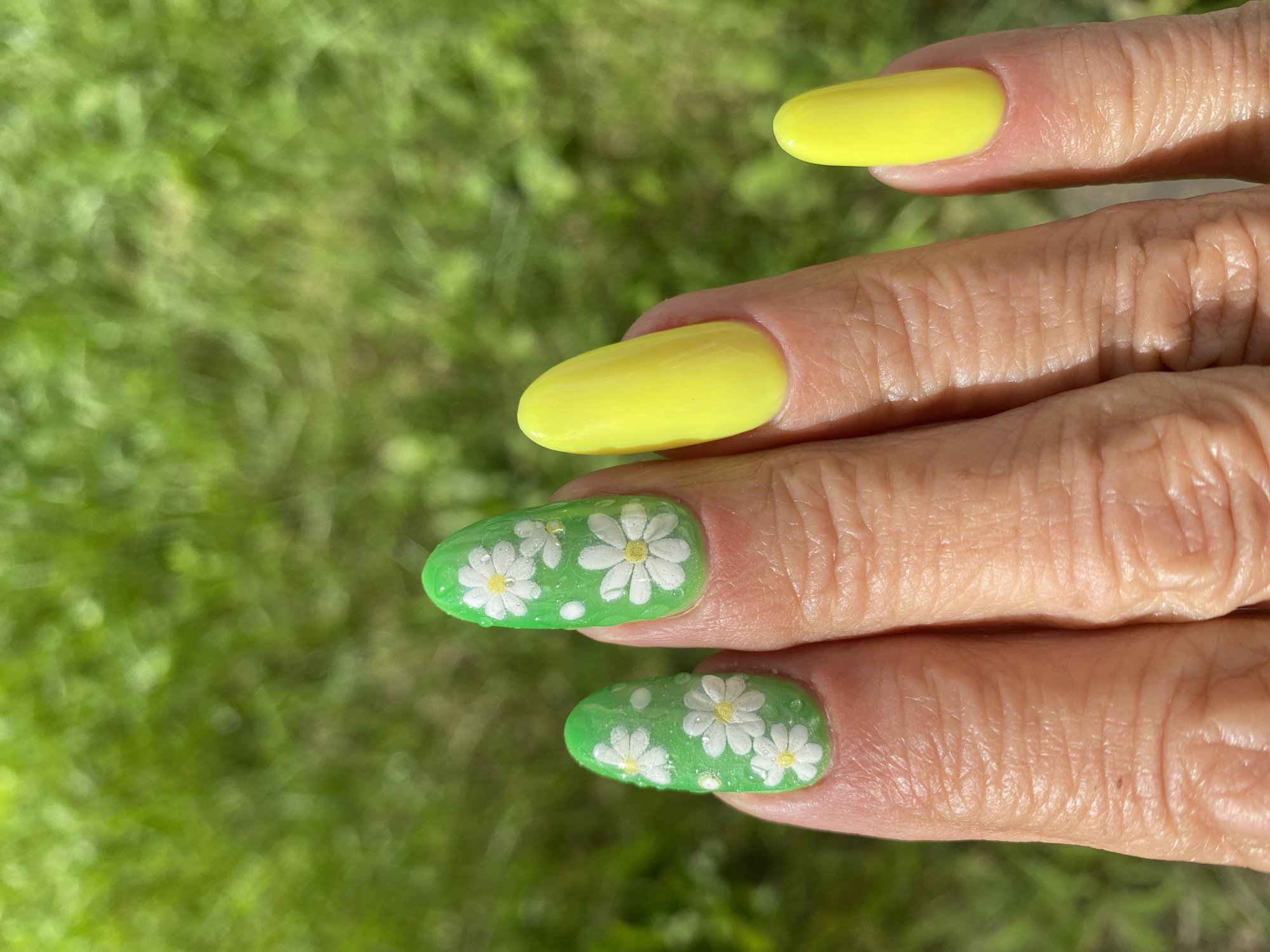 A close-up, eye-level shot shows a person's hand with fingernails decorated with a summer-themed manicure. The nails on the index and middle fingers are painted green and adorned with small white daisy designs and tiny clear embellishments. The ring and pinky fingers are painted a bright, solid yellow. The background is a blurred expanse of vibrant green grass, suggesting an outdoor setting. The image is taken on a sunny day. No specific location within Ivanteyevka, Russia, can be determined from the visual information provided.