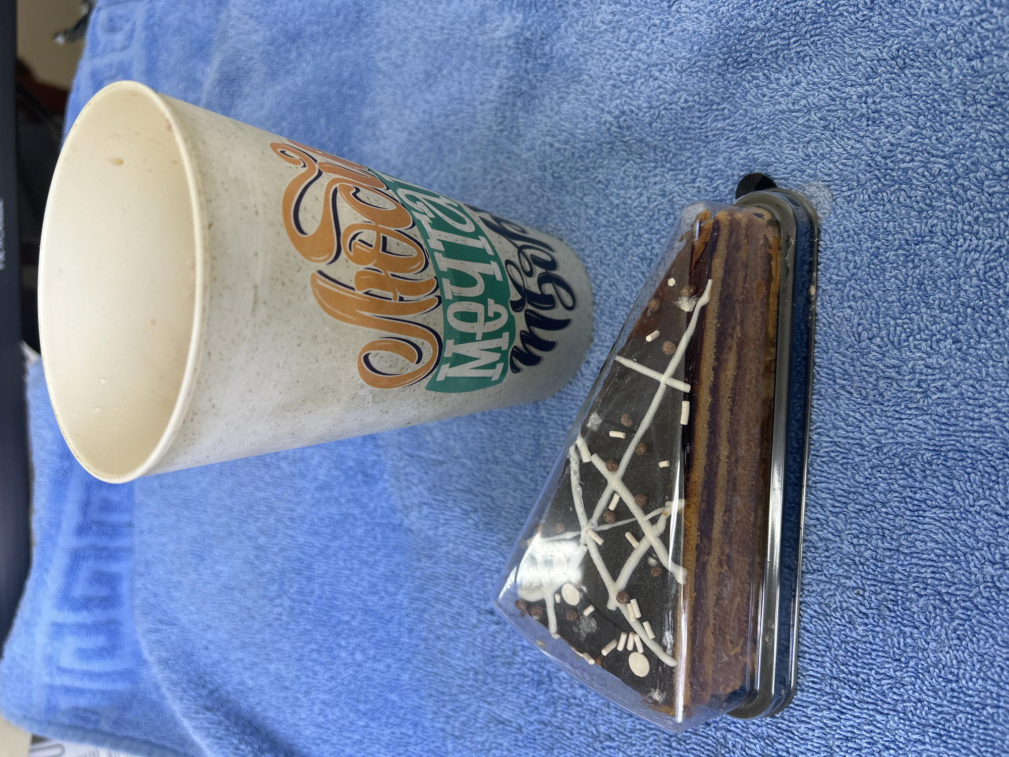 The image shows a cup and a piece of cake in a plastic container, lying on a blue towel. The cup is tan with orange, green, and black lettering. The cake is chocolate with white decorations on top and has multiple layers. The background is a light blue towel with a textured surface. I cannot determine any information about Фоки, Russia from the image.