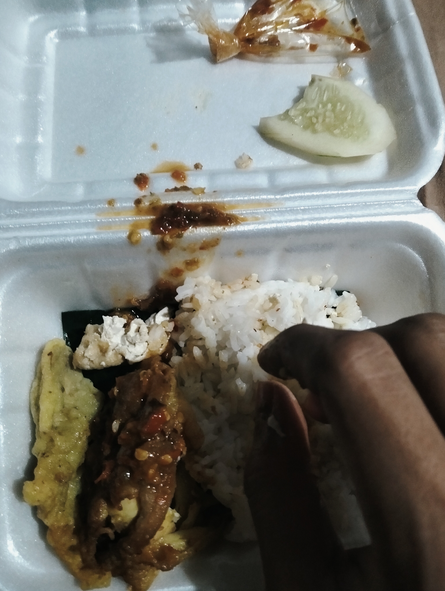 The image shows a close-up of a styrofoam takeout container filled with food, positioned within a city setting in Medan, Indonesia. The container holds white rice, a piece of fried food that appears to be tempeh or tofu, and a serving of meat in a dark sauce with visible chili flakes. There is also a slice of cucumber and some sauce residue in the upper part of the container. A person's hands are visible on the right side of the image, partially obscuring the rice as they appear to be interacting with the food. The overall lighting suggests it might be evening or indoors with artificial light. The background is blurred, but hints at an urban environment typical of Medan.
